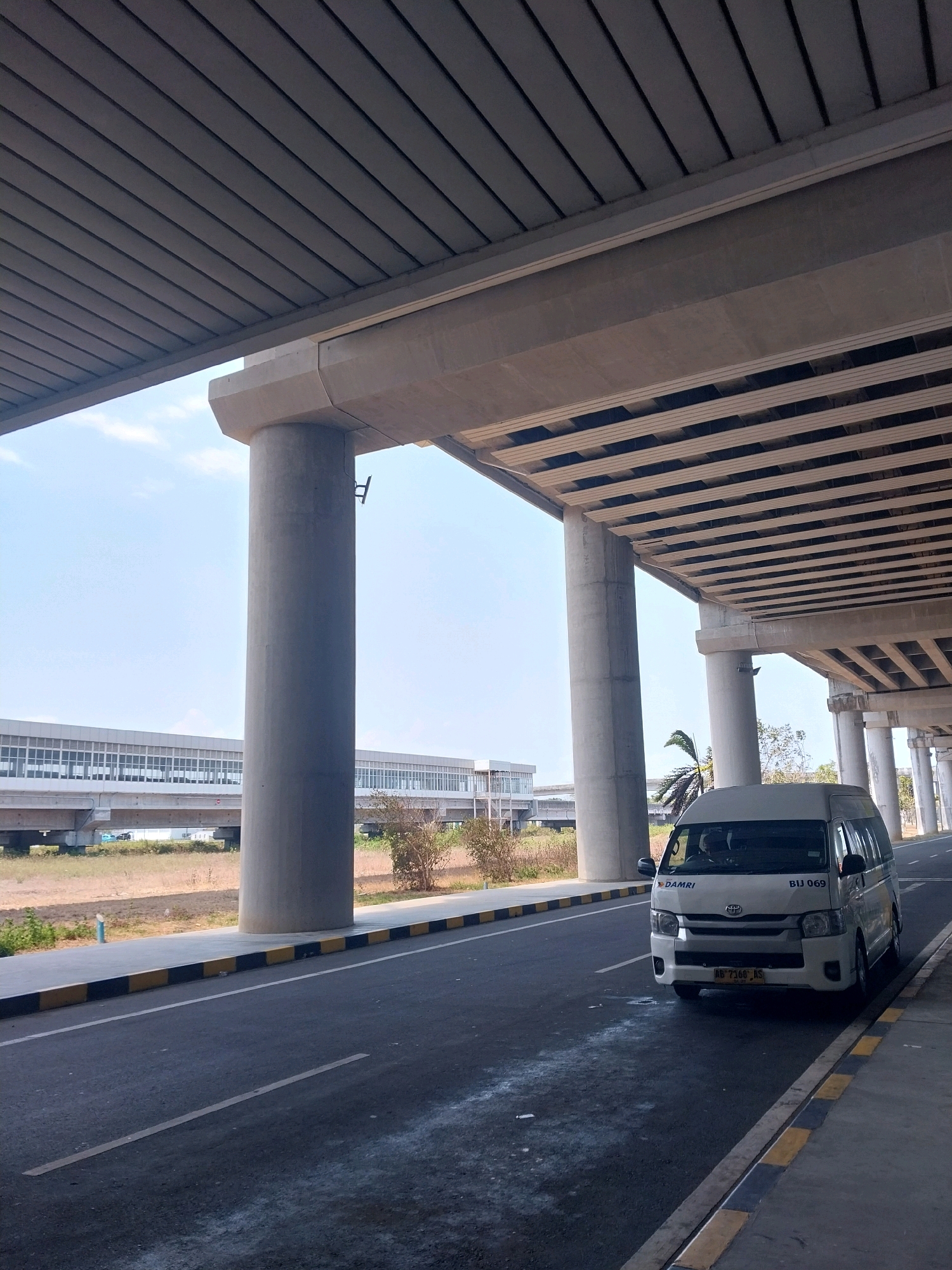 A white van with license plate AB 7160 AS is parked on the right side of an asphalt road, beneath an elevated structure supported by concrete pillars. The van has a logo with the text "DAMRI" and "BIJ 069" on its front. In the background, a long, modern building with numerous windows stretches across the landscape, likely a train station or airport terminal. The sky is clear and blue. The environment appears to be an urban or semi-urban setting in Palihan, Indonesia.