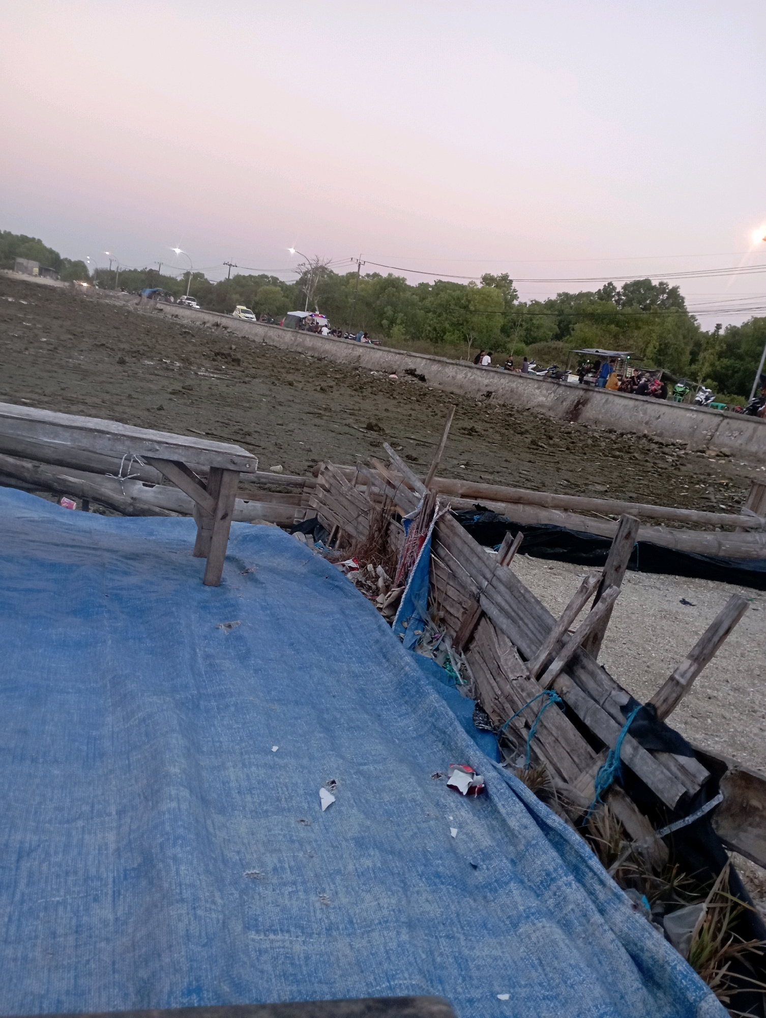 The image depicts a coastal area in Ngemboh, Indonesia, during the late afternoon or early evening. A muddy, exposed seabed is visible in the background, with a concrete embankment separating it from a road. Along the embankment, several vehicles, including cars and motorcycles, are parked, and groups of people are gathered. The sky is a pale pink and orange, indicating sunset. In the foreground, a blue tarp covers a surface, and a weathered wooden bench sits on top of it. To the right of the bench is a crude wooden fence or structure, made of salvaged materials. The overall scene suggests a casual gathering spot or a roadside rest area.