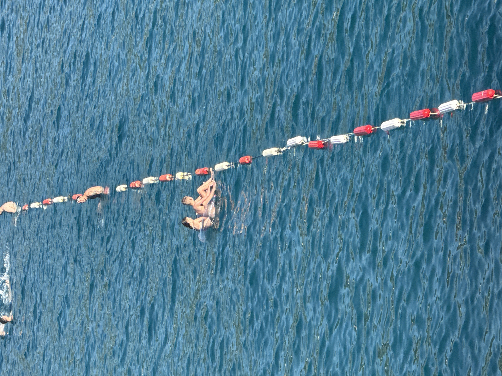 In Kamenovo, Montenegro, people swim in the bright blue sea, marked by a floating rope with red and white buoys. One group of swimmers is in the background, splashing in the water, while a man wearing a red cap is further down the rope. Two young women are in the middle, floating on an inflatable tube, facing each other. The rope of buoys extends diagonally from the top left to the bottom right of the frame, with the last buoy visible at the very bottom right corner of the image.
