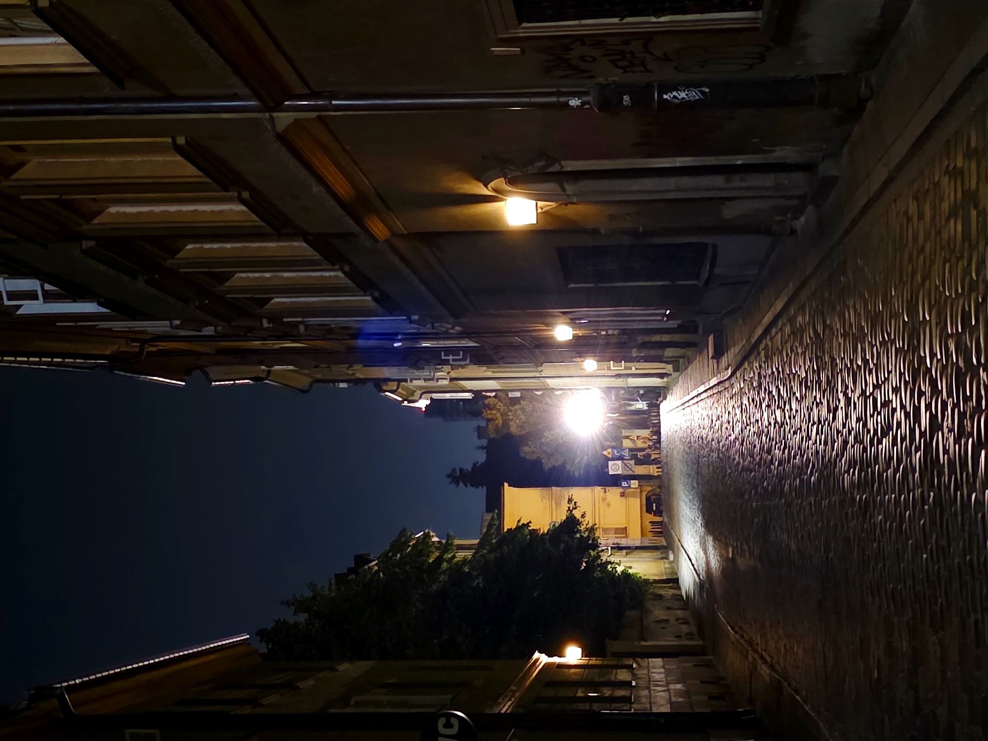The image depicts a cobblestone street in Warsaw, Poland, at night. The street is lined with old buildings, their facades illuminated by streetlights and light spilling from windows. Several lamps are visible on the buildings and on poles. Trees are present on the left side of the street, casting shadows. There are traffic signs and a car visible in the distance. The sky is dark and clear. Graffiti is visible on a lamppost on the right side of the image. The scene is devoid of any people or discernible activity.