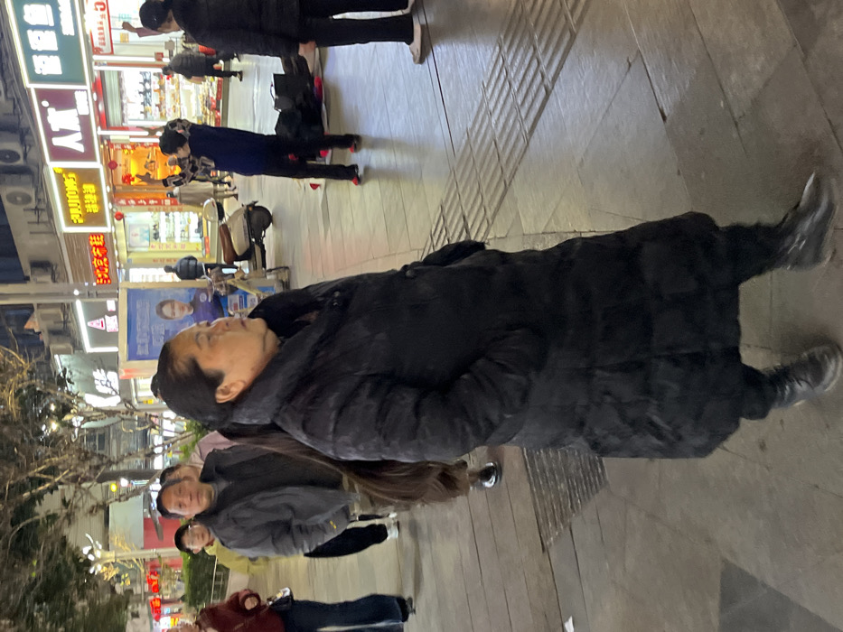The image depicts a nighttime or dusk scene on a pedestrian commercial street in Jiangjin, China. In the immediate foreground, a woman of East Asian descent is seen in profile, facing towards the right side of the frame. She wears a long, black, padded winter coat that reaches below her knees, black trousers, and black boots. Her dark hair is styled in a very long ponytail that extends down her back. Her hands are in her coat pockets, and her expression is neutral.
Behind the foreground subject and to her left, a man in a dark grey jacket and camouflage-patterned pants is observed walking, accompanied by a younger individual in a light yellow hoodie and dark pants. Further into the midground, a white scooter with a brown seat is parked on the paved sidewalk near a tall, blue advertising display board featuring a female face.
The background shows a row of brightly lit storefronts, consistent with a commercial district. Various signs with Chinese characters are visible, including a prominent red-on-gold sign reading "金金" (Jīnjīn) and another red sign reading "人杰" (Rénjié). Merchandise is displayed in the windows of these establishments. To the mid-right, a person in a blue top and dark pants is standing with their arms raised, appearing to gesture or interact with something out of frame. Several dark bags are positioned on the paved ground near this individual. Another person in dark clothing and light-colored shoes is partially visible on the far right. The ground is paved with rectangular concrete slabs, and a strip of tactile paving (grooved for the visually impaired) is present in the lower right section of the image. Artificial lighting from storefronts and overhead sources illuminates the entire scene.