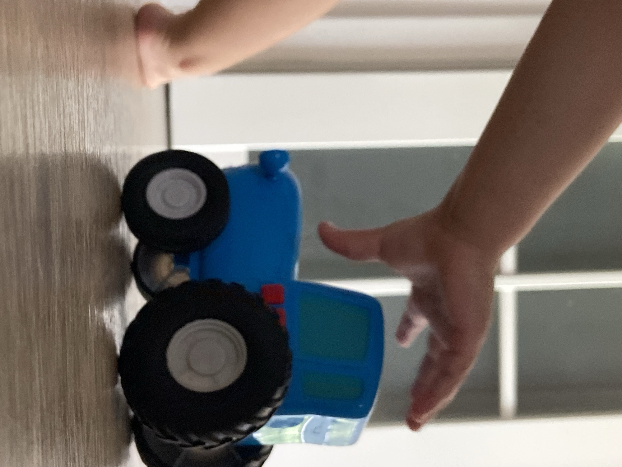 The image displays an indoor scene, likely in Tyumen, Russia, rotated 90 degrees counter-clockwise. A blue toy truck, possibly a tractor, is lying on its side in the center of the frame, featuring black tires with grey hubs and accents of red and teal. One hand, presumably a child's, is visible at the top-left, resting with fingers lightly curled on a light brown, wood-grained surface. Another hand, also likely a child's, is positioned on the right side of the frame, fingers slightly extended as if reaching towards or interacting with the toy truck. The background on the right consists of light grey and white rectangular panels or blinds, suggesting a window or a paneled wall, which indicates a bright, daytime setting. No discernible text is visible.