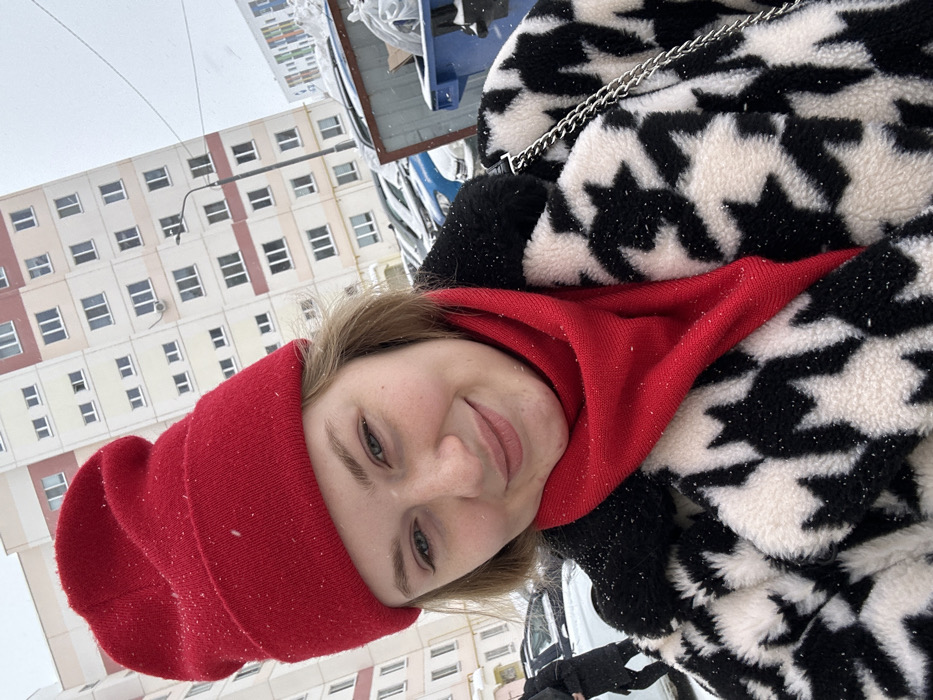This image captures a winter street scene in the city of Nizhnekamsk, Russia, under conditions of light, active snowfall. The primary subject is a young adult female, positioned for a selfie, smiling slightly and looking directly at the camera. She has light brown hair and is dressed in a bright red knit beanie and a matching red scarf or turtleneck. Her outer garment is a black and white geometric-patterned (likely houndstooth) fuzzy coat, with visible snowflakes dusting her clothing. A silver-colored chain, possibly from a shoulder bag, is visible on her right side.
In the background, a row of parked vehicles, partially covered in snow, lines what appears to be a residential street or parking area. A black streetlight with a single luminaire stands behind the cars. To the right, a large, grey industrial dumpster and a smaller blue recycling bin, both accumulating snow, are present. Dominating the left background is a multi-story apartment building with a light beige or pale yellow facade and reddish-orange window accents, featuring numerous rectangular windows and some external air conditioning units. Further right, a portion of another multi-story building with colorful facade elements (greens, blues, reds) is visible. On the far left of the frame, the lower body of an individual in dark clothing is partially visible, walking away from the camera. The sky appears overcast, contributing to the winter atmosphere.