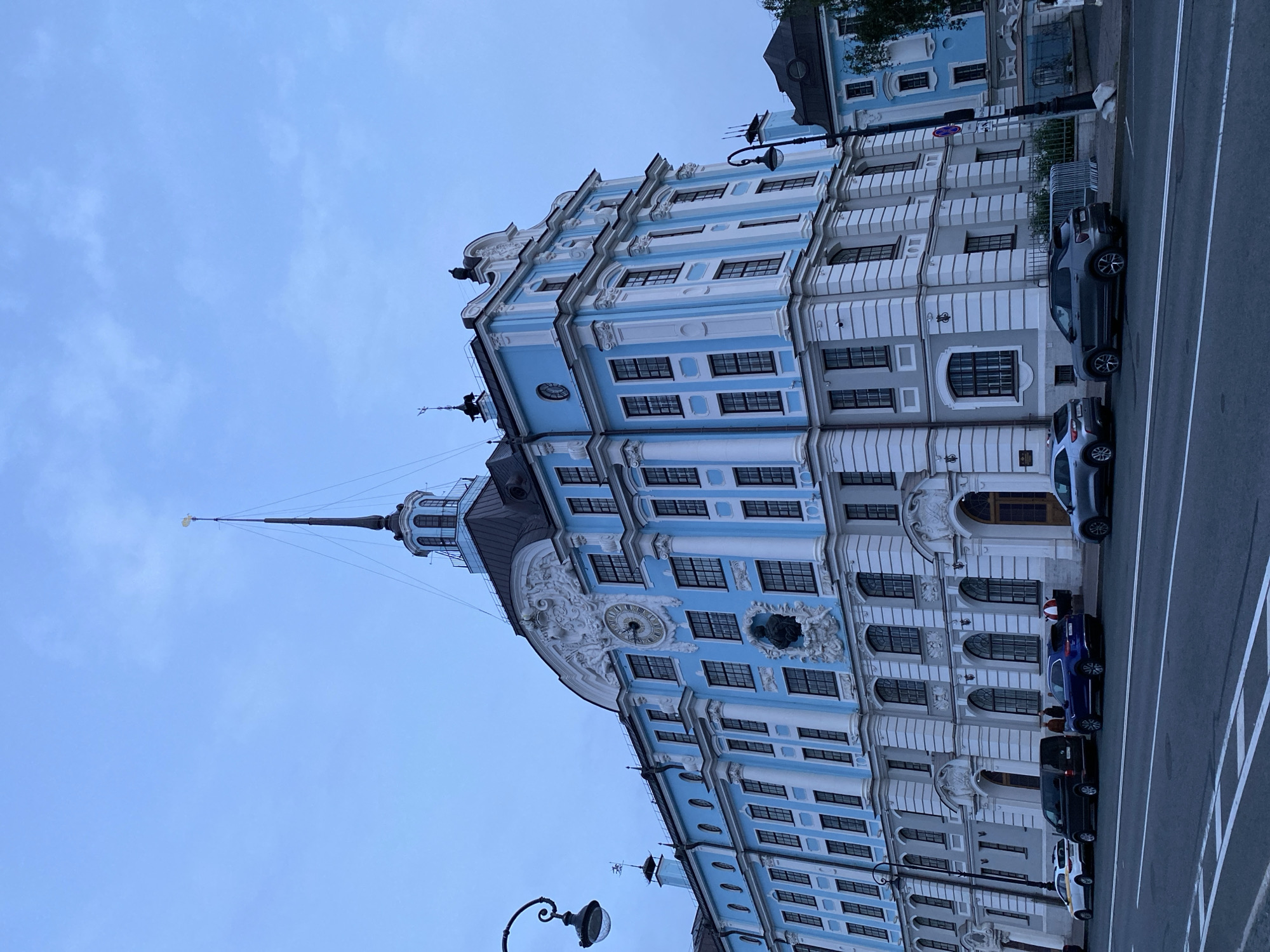 The image depicts a grand, historic building with a striking blue and white facade, characteristic of Baroque architecture. The building features multiple stories, adorned with intricate decorative elements, columns, and numerous windows with dark frames. A prominent spire crowns one section of the building, adding to its majestic appearance. In front of the building, cars are parked along the side of a street. The sky is overcast, suggesting a cloudy day. The overall scene evokes a sense of historical significance and urban grandeur, typical of a city like Saint Petersburg, Russia. The time of day appears to be late afternoon or early evening, given the muted light. No people are clearly discernible, and no visible text is present in the image.