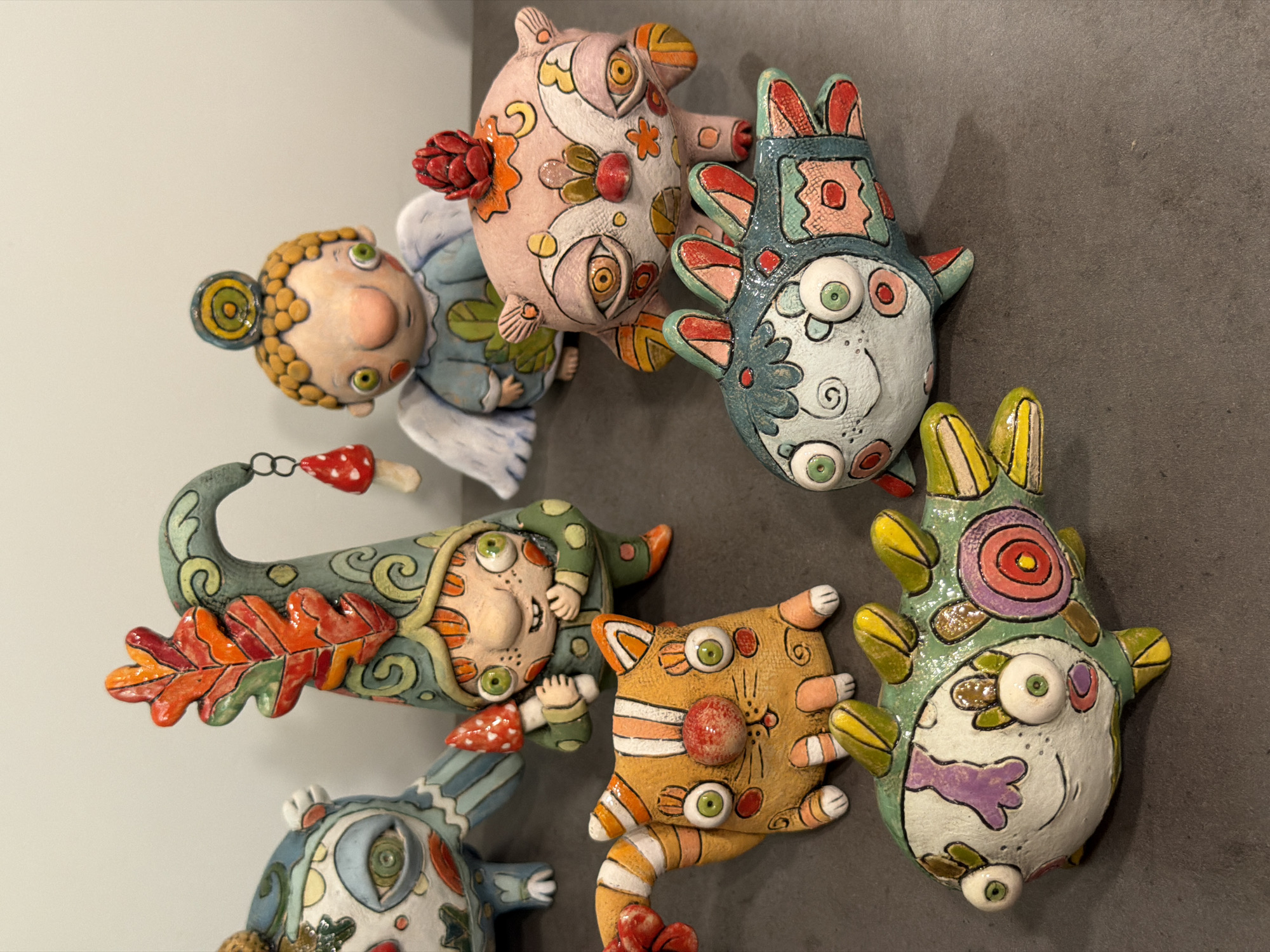This is a close-up, eye-level studio photograph of a collection of six whimsical, handmade ceramic figurines arranged on a dark grey, matte surface. The figurines are brightly colored and feature stylized, almost cartoonish designs with large, expressive eyes.

From left to right, the figurines are:
1. A blue and white creature with large, detailed eyes, striped legs, and a patterned body.
2. A gnome-like figure wearing a green, patterned cap adorned with an orange and red oak leaf. The gnome has freckles, big green eyes, and is holding a small red and white mushroom.
3. An orange cat with a striped tail, large green eyes, and a red nose. It has white stripes on its body and paws.
4. A small angelic figure with blonde curly hair, green eyes, and a blue garment with leaf motifs. It has white wings and a red bud-like ornament on its head.
5. A pinkish creature with large, multifaceted eyes and red, orange, and yellow floral or organic patterns across its body. It has small, wing-like appendages and a red bud on its head.
6. A green and teal fish-like creature with large, round eyes, colorful scales, and yellow and red fin-like protrusions.

The lighting appears to be even and bright, illuminating the details of the figurines. The background is a plain, light grey wall, suggesting an indoor studio or display setting. There is no indication of the specific location within the city of Askat, Russia, nor any environmental context outside of the immediate display area. The image captures a static arrangement of these art objects.