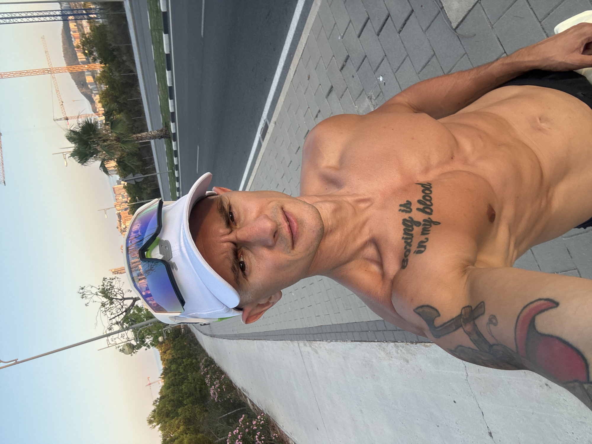 A shirtless man with short dark hair, wearing a white baseball cap and blue-lensed sunglasses, stands on a paved sidewalk next to a road in Finestrat, Spain. He has visible abs and tattoos on his left arm and chest. The tattoo on his chest reads "boxing is in my blood." He is taking a selfie with a slight frown. The background shows a street with a white line, a palm tree, and buildings on a hill with construction cranes visible. The sky is clear and light blue, suggesting it is daytime.