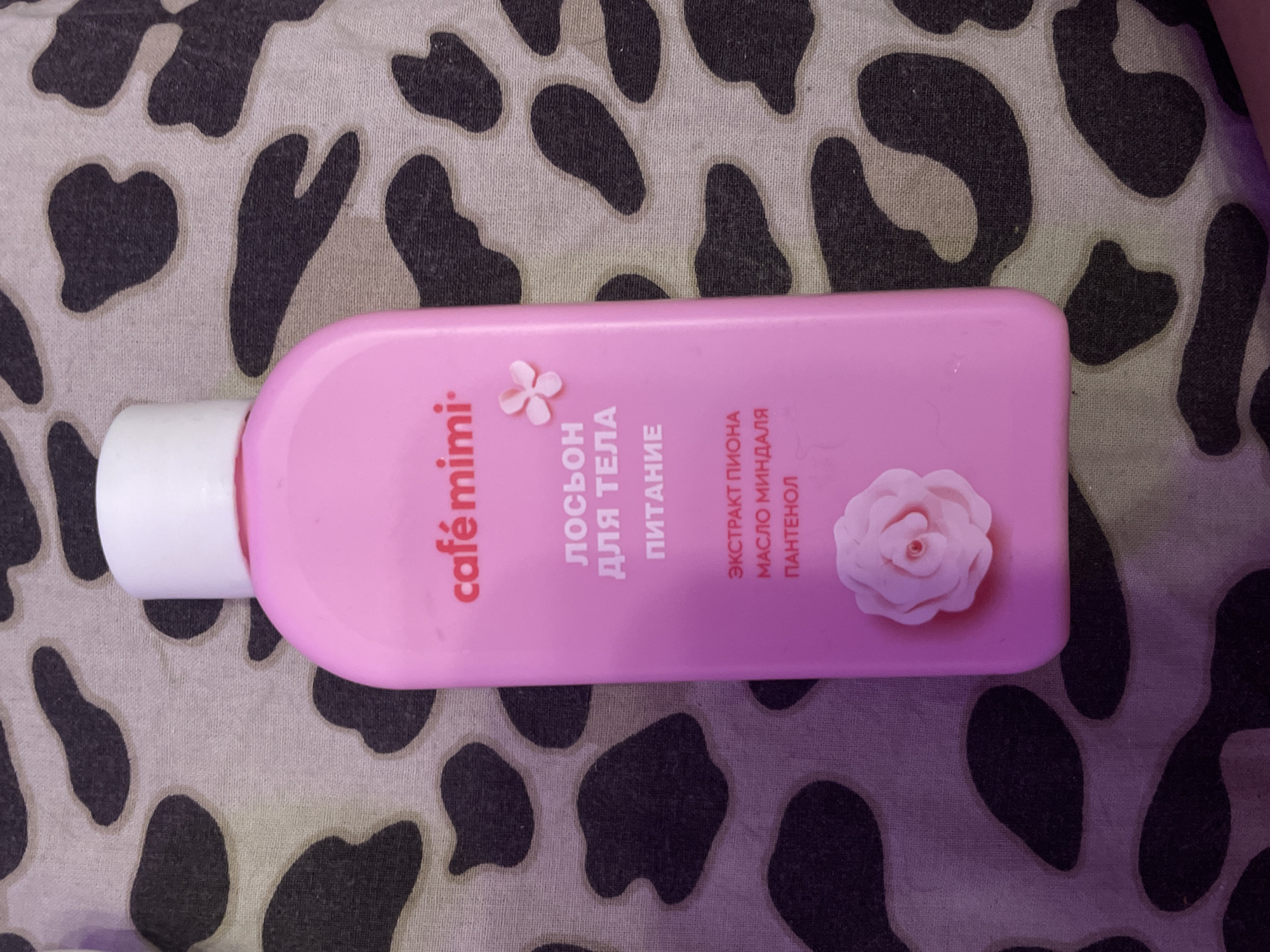 A pink bottle of "Café mimi" body lotion with a white cap is placed on a patterned fabric. The fabric has a spotted design, reminiscent of cow print, in black and grey on a light background. The lotion bottle features text in Russian, including "ЛОСЬОН ДЛЯ ТЕЛА ПИТАНИЕ" (Body Lotion Nourishing), and lists ingredients such as "ЭКСТРАКТ ПИОНА" (Peony Extract), "МАСЛО МИНДАЛЯ" (Almond Oil), and "ПАНТЕНОЛ" (Panthenol). A small pink floral design is visible on the bottle above the text, and a larger pink rose embellishment is present on the lower part of the bottle. The image is taken from a top-down perspective. The background pattern suggests the item is likely at a domestic setting within Shcheglovo, Russia.