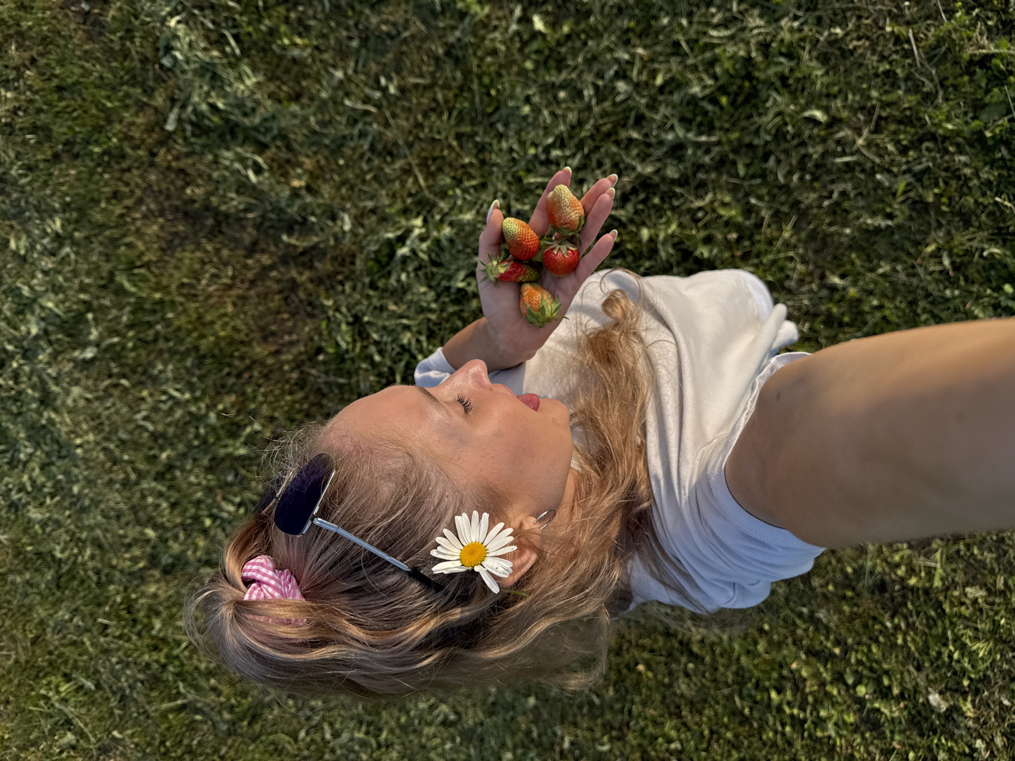 The image shows a woman lying on grass in Nizhnyaya Chermoda, Russia. She is holding a handful of strawberries, with several more visible in her palm. She has a daisy tucked behind her ear and is wearing sunglasses. Her hair is pulled back with a pink and white checkered scrunchie. The scene appears to be set during the daytime, with natural lighting casting shadows on the grass. The woman has a relaxed expression, with her head tilted back and her eyes closed. The overall mood is one of leisure and enjoyment of nature. There is no visible text in the image.