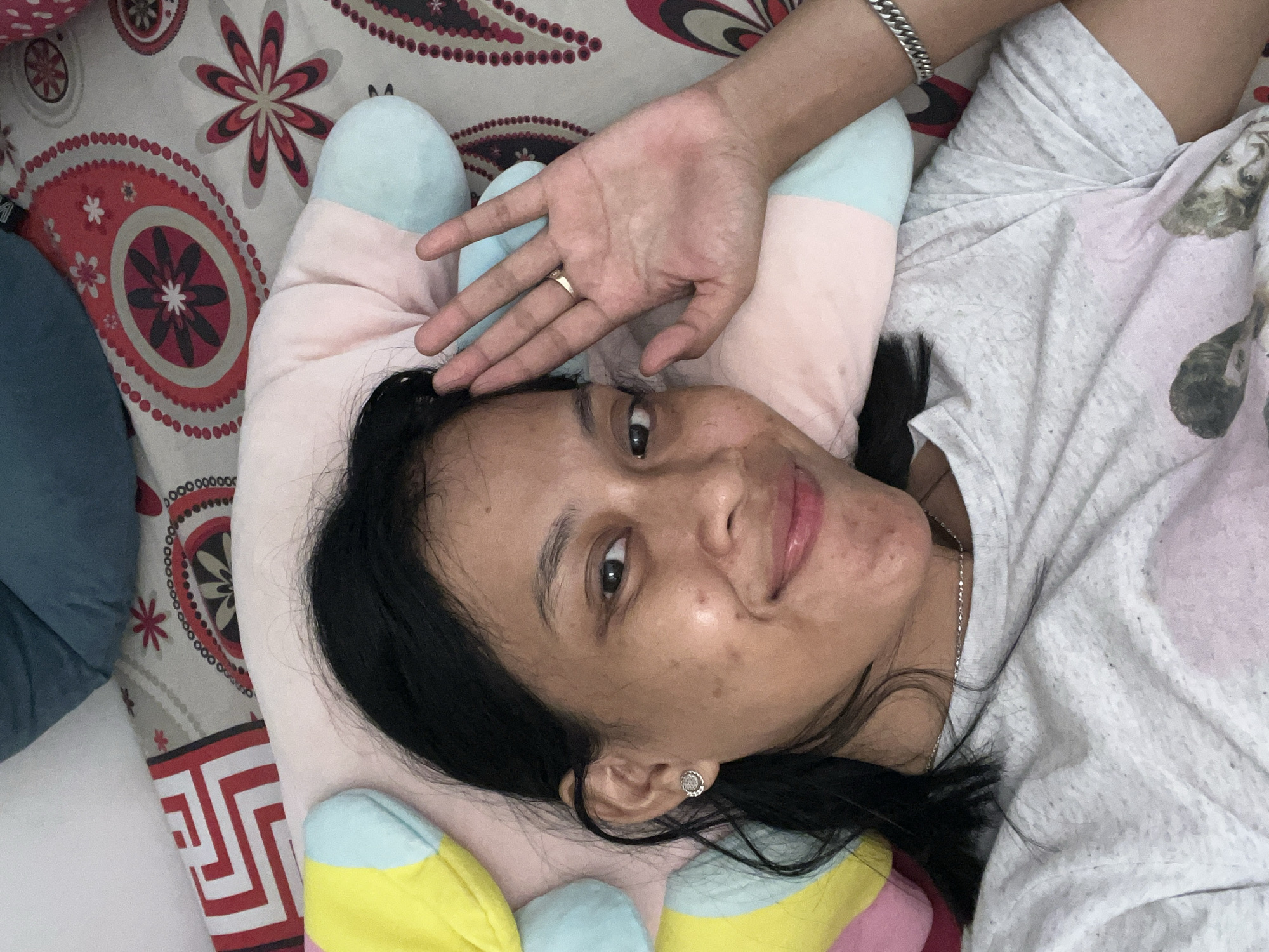 A selfie of a woman lying down on a bed with colorful pillows and patterned sheets. She is looking at the camera and waving with her right hand. She is wearing a grey t-shirt and has a ring on her right ring finger and a bracelet on her right wrist. The woman appears to be indoors in a bedroom in Kecamatan Pati, Indonesia.