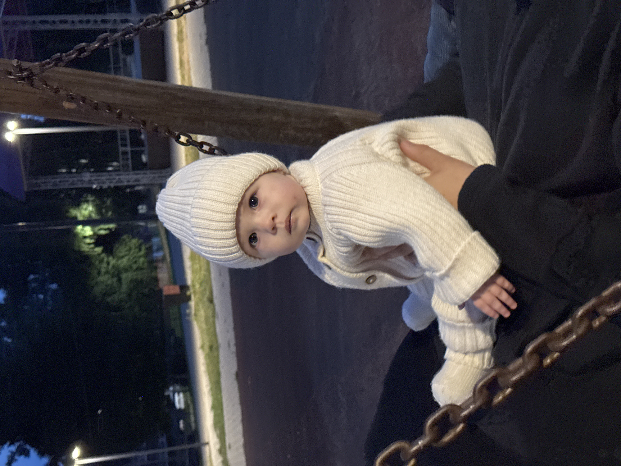 A baby wearing a white knitted hat and sweater is sitting on someone's lap outdoors in Noviy, Russia. The baby is facing forward, with a neutral expression. The background is dark and out of focus, but appears to be a park or playground area with some lighting and trees. A chain from a swing set is visible in the foreground.