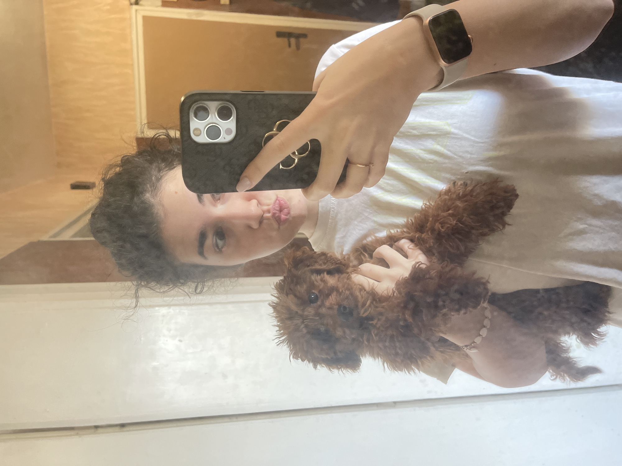 A woman is taking a selfie in a mirror, holding a brown poodle. She is wearing a white t-shirt, a watch on her left wrist, and a ring on her left hand. The phone she is using has a dark-colored case with a flower-shaped grip. The background includes a light-colored wall and a wooden doorframe. The location is not evident in the image to determine it to be in Sofia, Bulgaria.