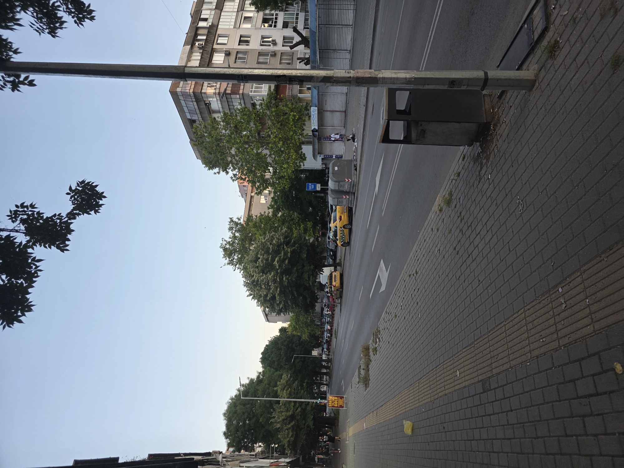 The image depicts a street scene in Varna, Bulgaria, possibly along a main thoroughfare. The perspective is from the sidewalk looking down the street. On the left side of the street, there are buildings with some trees in front of them. A yellow sign with Cyrillic text is visible on the sidewalk. Further down, a line of yellow taxis is parked. The street itself has multiple lanes of traffic and is lined with trees on the right side. In the background, there are apartment buildings and some shops with roll-up shutters. A metal pole, likely a lamppost, is prominent in the foreground on the right, with a public trash receptacle next to it. The sky is clear and blue, suggesting it is daytime. There are a few people walking on the sidewalk on the left, and one person standing near the shops on the right. The overall impression is of an urban environment during daylight hours.
