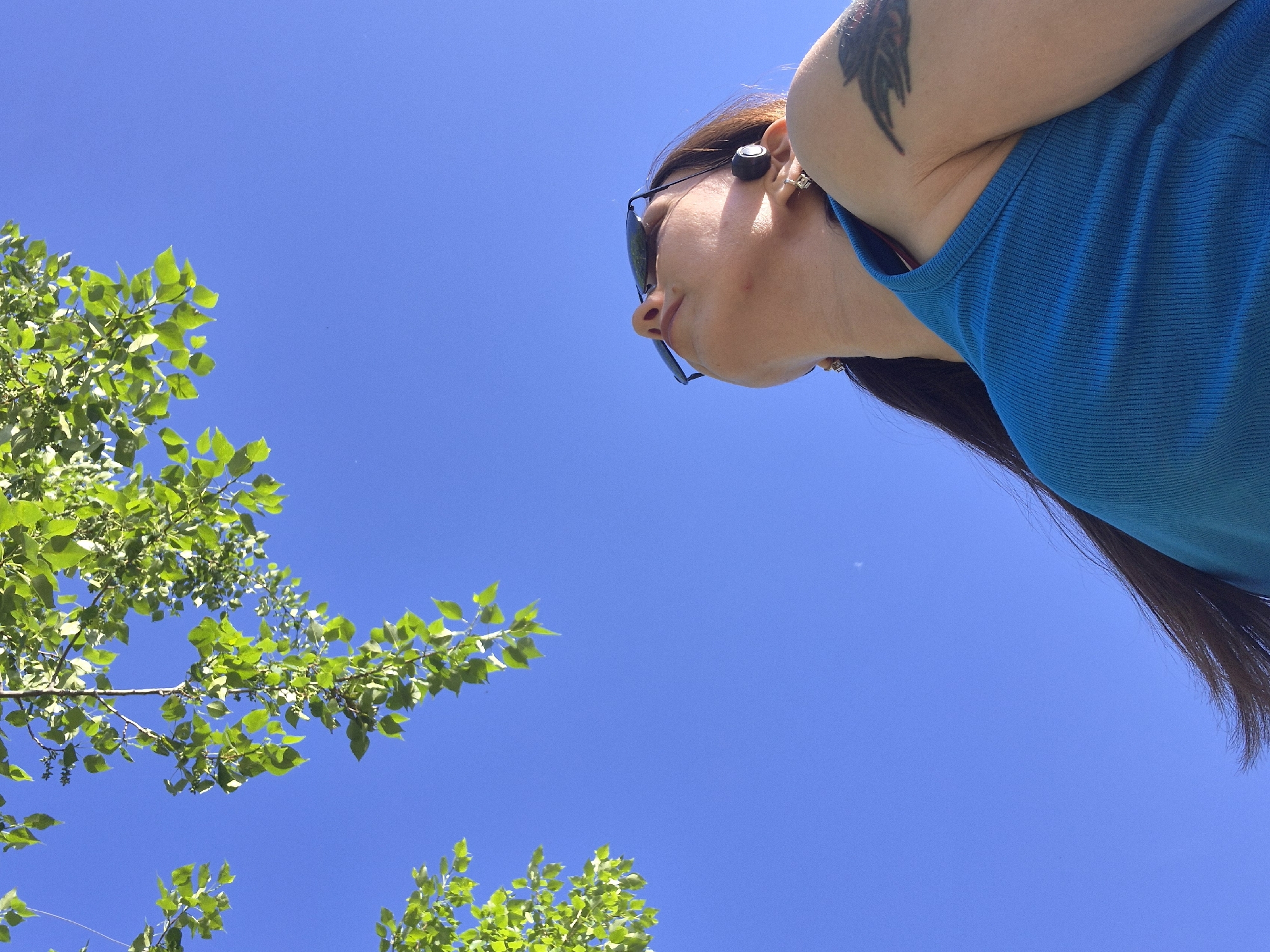 A low-angle shot depicts a woman wearing sunglasses and a blue tank top, with a tattoo on her upper arm. She is wearing a black device in her ear. Her gaze is directed towards the lower left corner of the frame, where branches of a tree with green leaves are visible against a clear, bright blue sky. The location appears to be outdoors, potentially in a city park or a similar open space.
