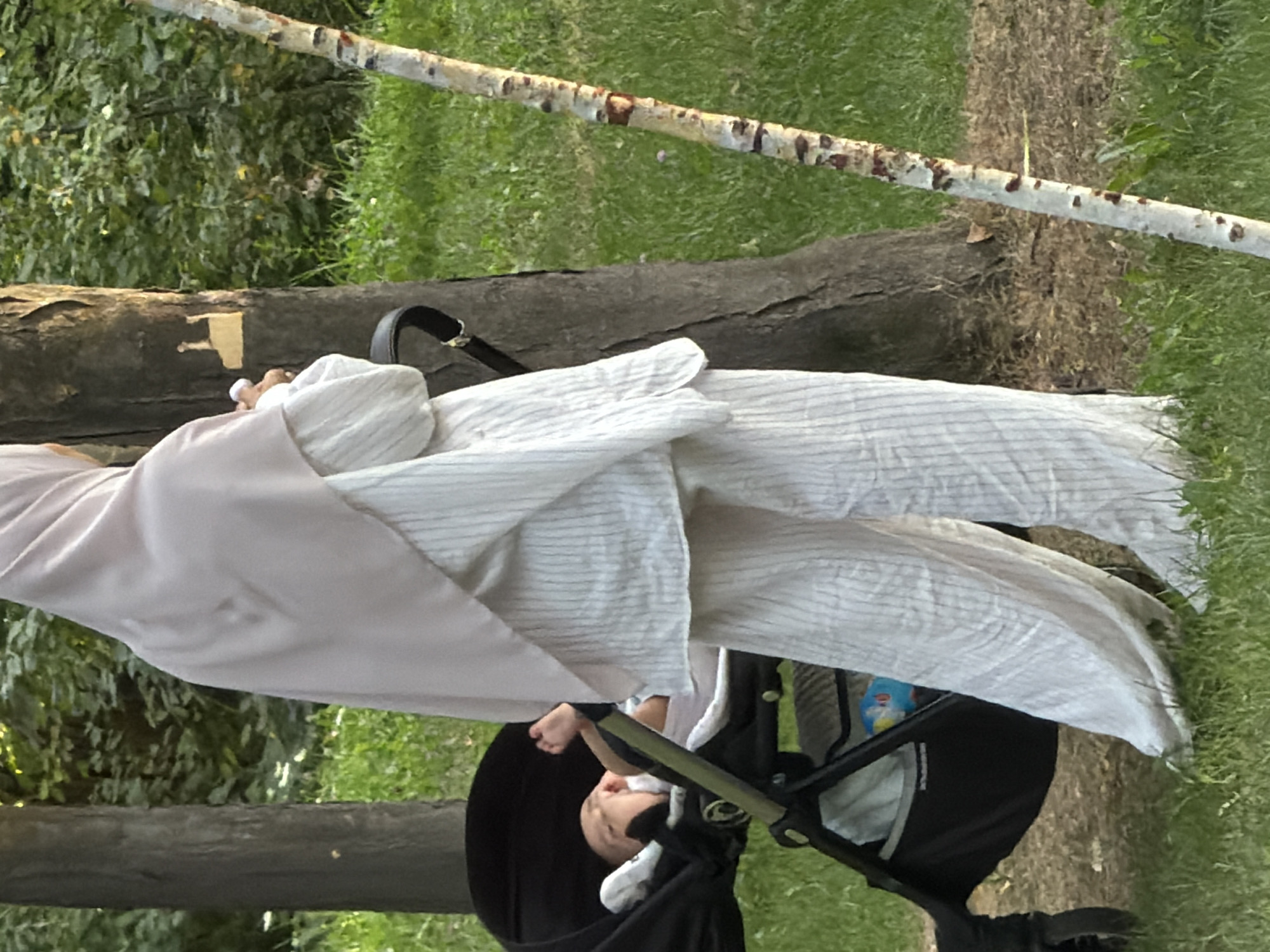 An individual wearing a pale pink hijab and a white striped outfit stands in a park, partially obscuring a baby in a black stroller. The baby, lying down, is facing the camera and appears to be looking up. The individual is holding a small white object in their right hand. The scene is set amongst trees and grassy terrain, with a thin, white-barked tree trunk to the right of the frame. The background consists of green foliage and dappled sunlight.