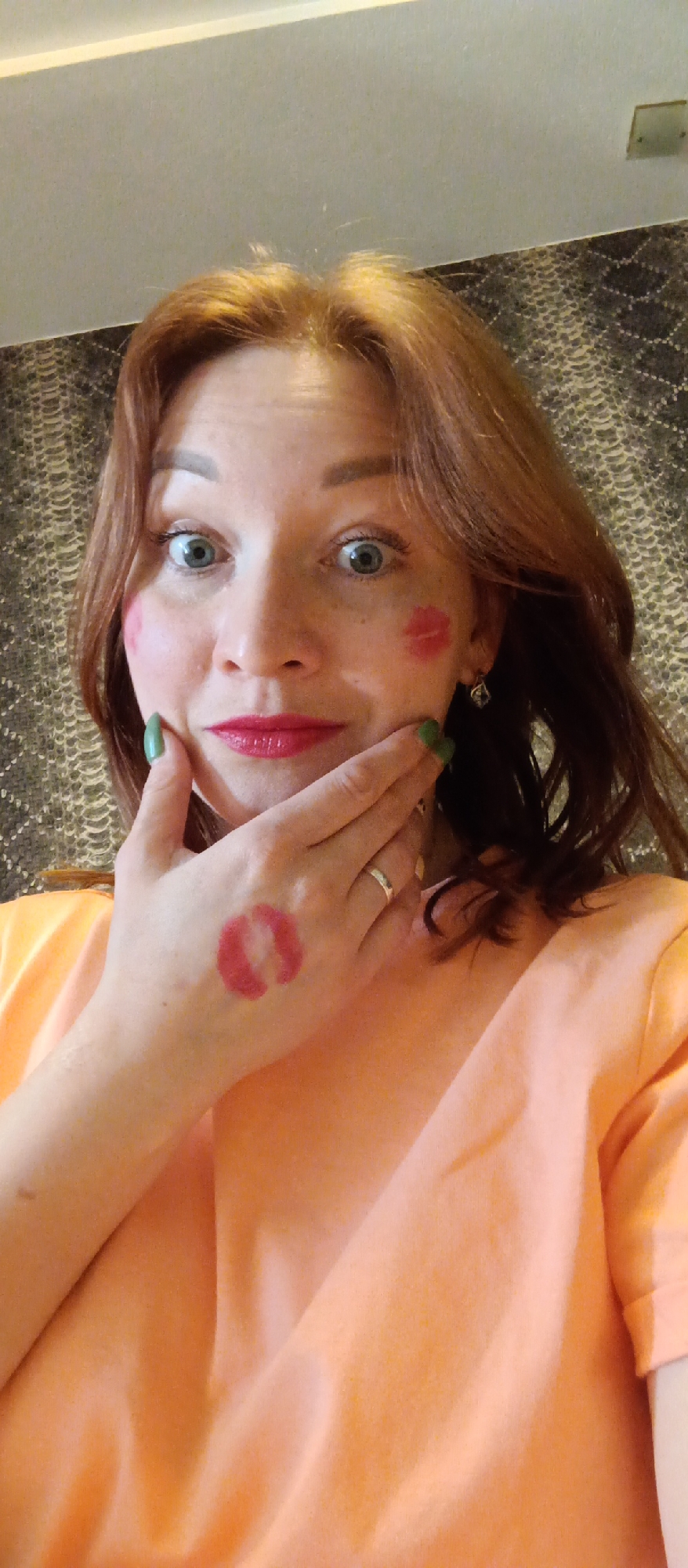 A person with light skin and reddish-brown hair poses for a selfie. They are wearing an orange shirt. A lipstick kiss mark is visible on their left cheek and the back of their left hand. Their fingernails are painted green. They are wearing a silver ring on their left ring finger. The background is a textured wall. The location is Unknown, Unknown.