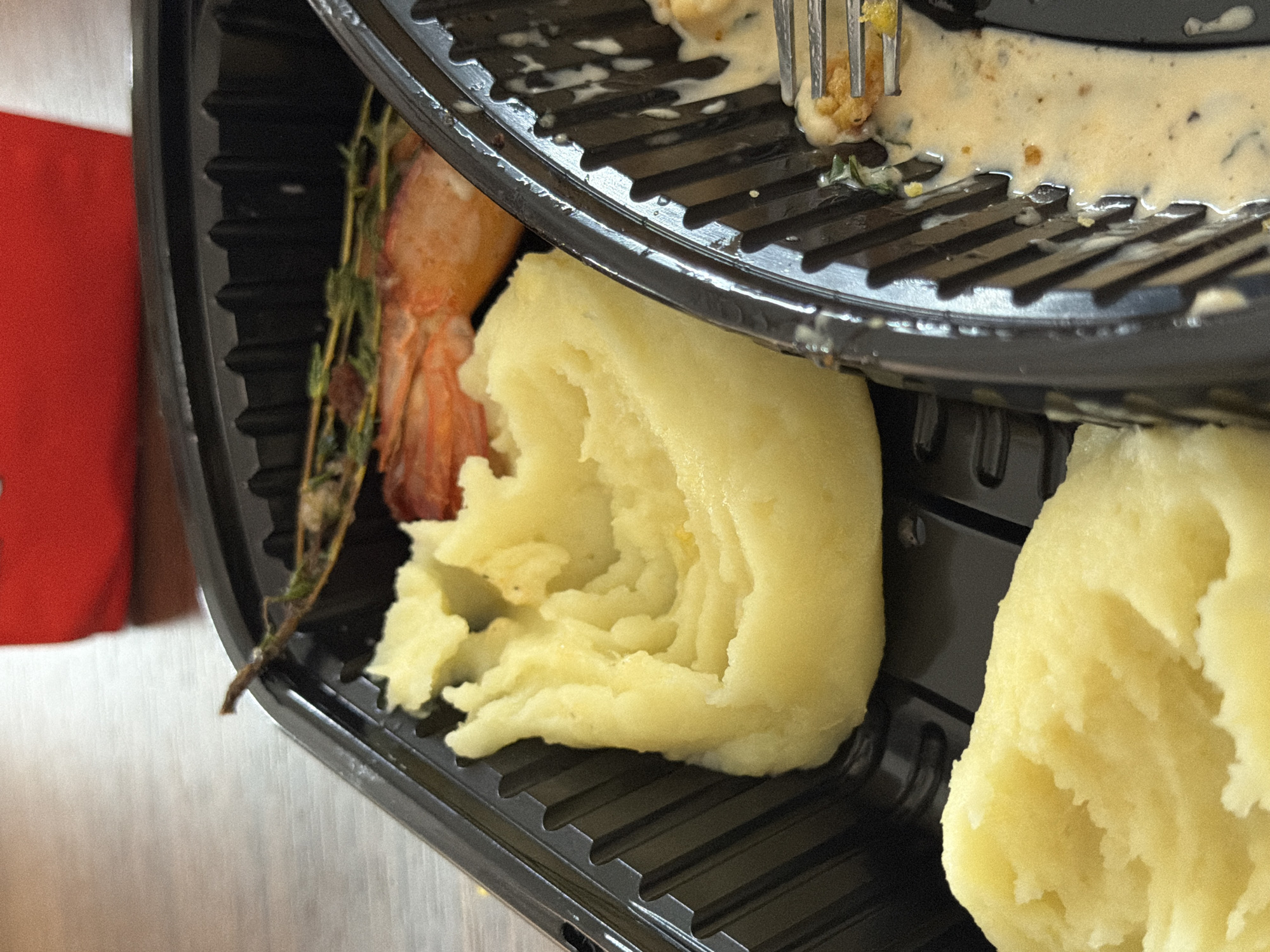 The image shows a close-up of a take-out container with food. The container is black plastic and divided into sections. In one section, there is a portion of mashed potatoes, which appears to be creamy and yellow. In another section, a single shrimp is visible, along with a sprig of herbs. A fork is seen in another section of the container, with some food and creamy sauce. In the background, a red object is partially visible, and a wooden table surface is also apparent. There is no indication of the city of Veliko Tarnovo, Bulgaria.