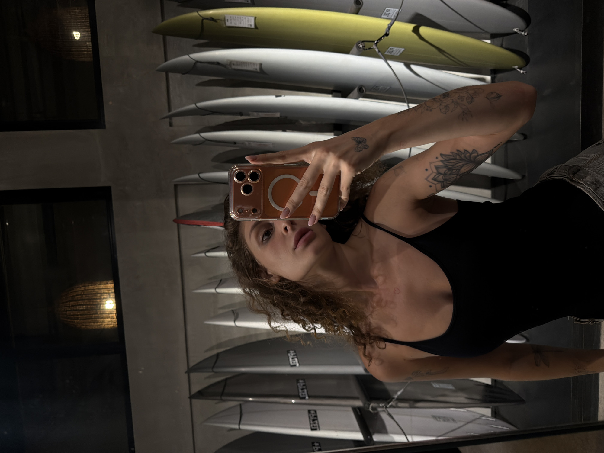 A young woman with curly brown hair takes a mirror selfie in a shop in North Kuta, Indonesia. She is wearing a black tank top and denim shorts. In the background, several surfboards are displayed vertically on racks. The lighting suggests it is evening or indoors.
