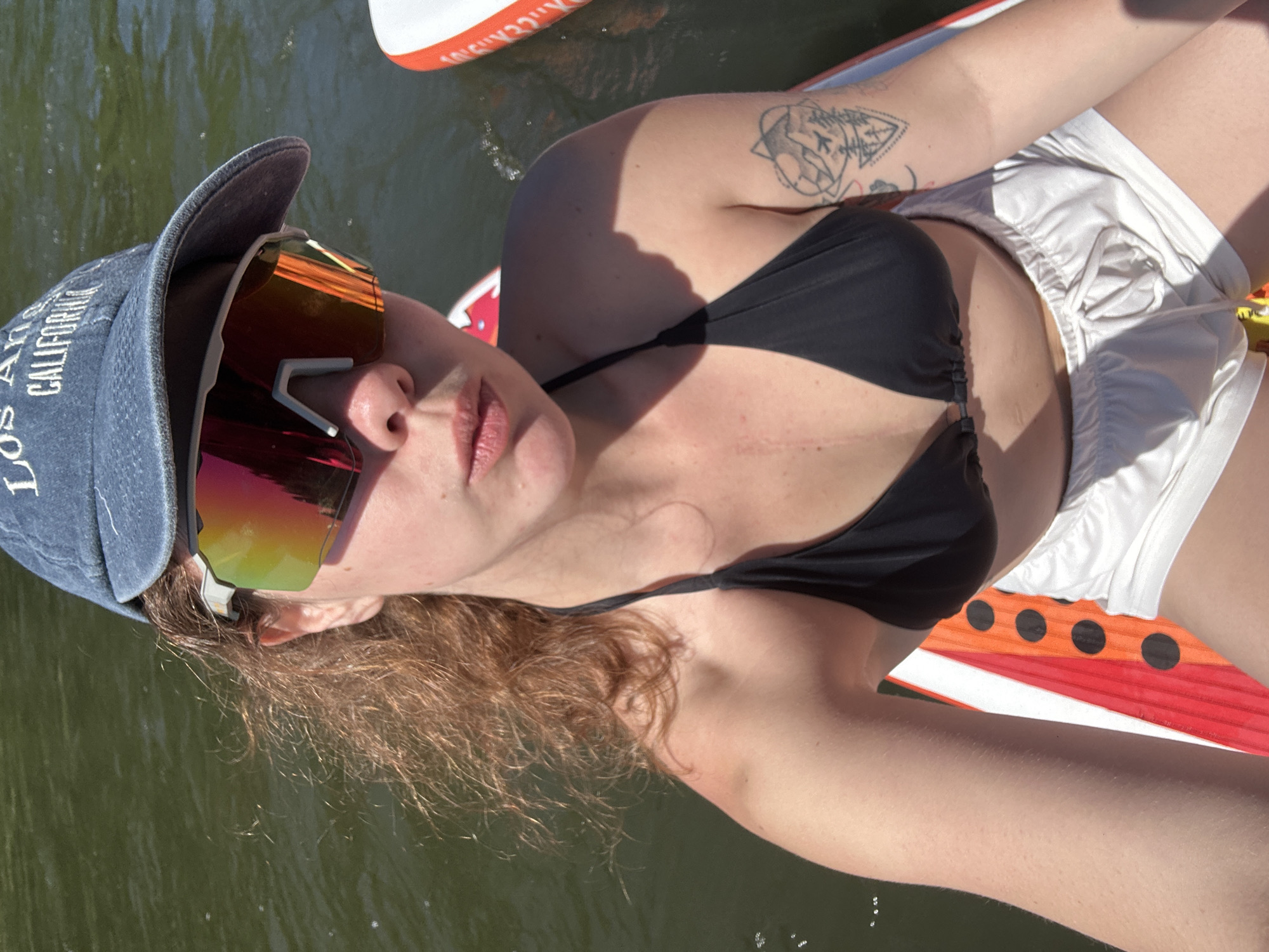 The image shows a young woman with reddish-brown curly hair wearing a black bikini top, white shorts, a denim baseball cap with "Los Angeles California" embroidered on it, and reflective sunglasses. She is sitting on an inflatable paddleboard, which is partially submerged in dark green water. The paddleboard has orange and white stripes with black circles. The woman is looking at the camera with her mouth slightly open. There is a tattoo of mountains and an airplane on her upper left arm. The scene is outdoors, likely on a river or lake, given the water and the inflatable paddleboard. The lighting suggests it is daytime and sunny. The location is identified as Ust-Mana, Russia, though the image itself does not contain any explicit visual cues to confirm this location.