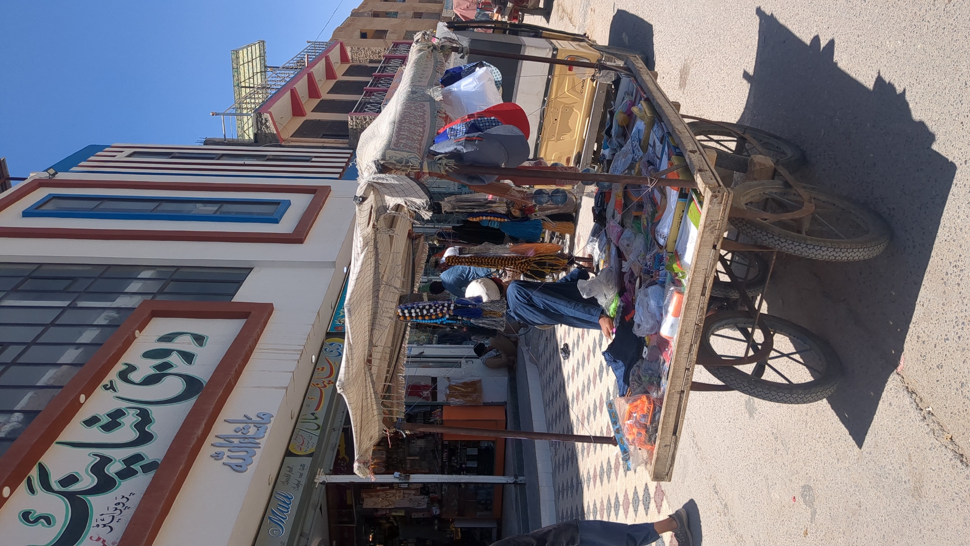 The image shows a street scene in Quetta City Tehsil, Pakistan. A vendor is selling goods from a pushcart. The cart is filled with various items, including beads, clothes, and toys. The vendor is standing behind the cart, interacting with customers. The cart is parked in front of a building with a storefront displaying signs in Urdu. The street is paved and partially shaded by the cart and nearby buildings. The sky is clear and blue.