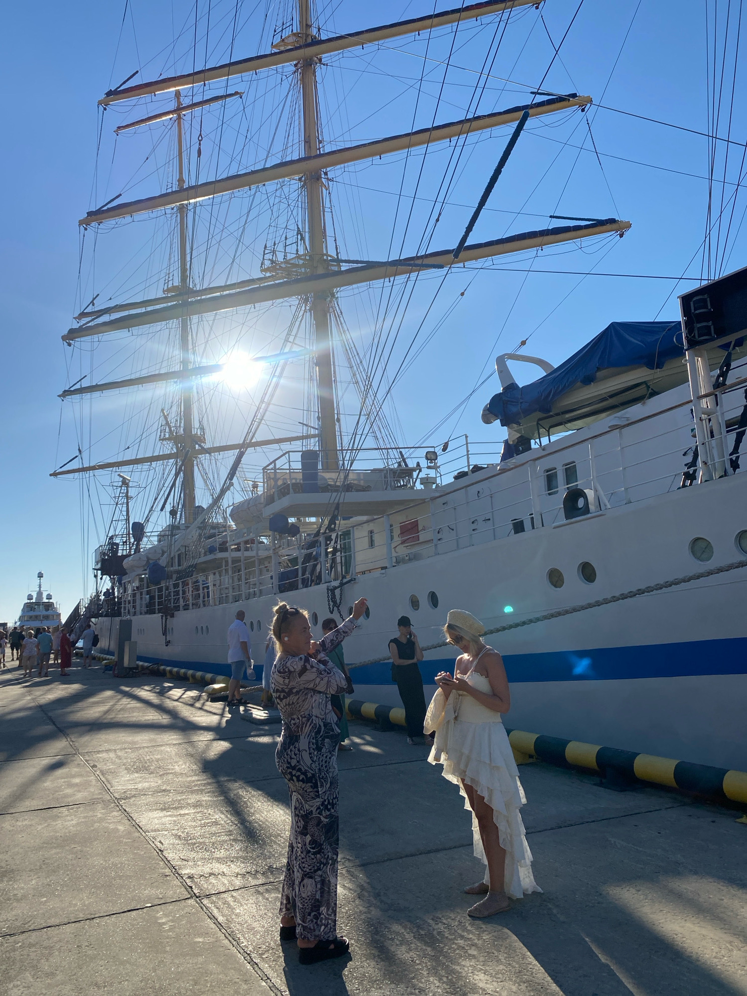 The image depicts a sunny day at a seaport in Sochi, Russia. A large, white sailing ship with multiple masts dominates the background. In the foreground, two women are visible. One woman, on the left, is wearing a patterned jumpsuit and is holding up a phone, appearing to take a picture. The second woman, on the right, is dressed in a white, layered dress and a straw hat, and is looking down at her phone. Several other people are walking along the dock in the background. The dock itself is made of concrete, with yellow and black bollards along the edge of the water. The sun is bright, casting long shadows on the dock. Another smaller boat is docked in the distance to the left.