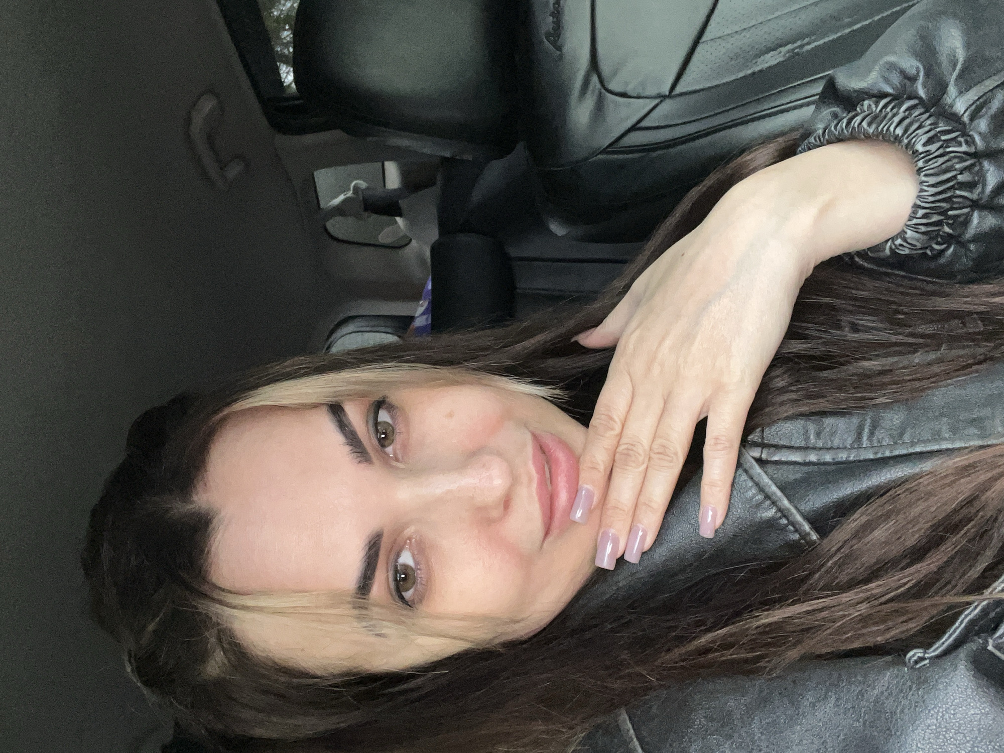 A young woman is shown in a medium close-up, angled diagonally, suggesting a selfie taken inside a car. She has long, dark brown hair with a prominent blonde streak framing the left side of her face. Her fair skin is accented by dark, well-defined eyebrows, brown eyes, and lips covered in a light pink gloss, forming a gentle smile. She wears a black jacket, appearing to be leather or a similar material, with a distinctive ruched or elasticated cuff visible on her right arm. Her left hand is placed near her chin, displaying square-tipped nails with a light pink or translucent purple polish. The background reveals the dark interior of a vehicle, including a dark car seat, a portion of the car's ceiling, and a grey grab handle. The lighting is soft and natural, indicating it is daytime. She appears relaxed and is looking directly at the camera. There is no visible text in the image.