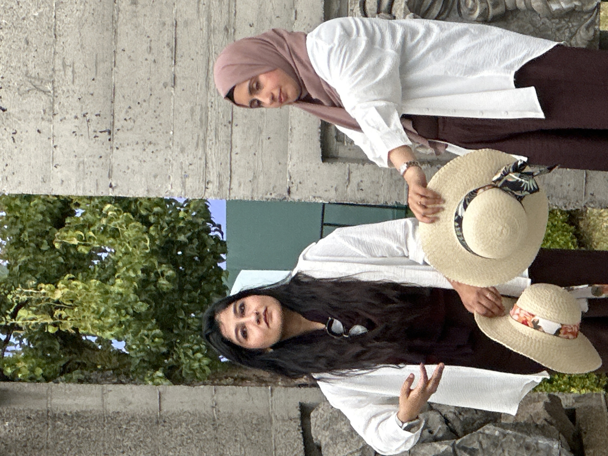 Two women are posing outdoors in front of a grey concrete wall. The woman on the left has long, dark hair and is wearing a white open shirt over a dark brown dress. She is holding a straw hat decorated with a floral ribbon in her left hand, and her right hand is gesturing. The woman on the right is wearing a pink hijab and a white open shirt over dark brown trousers. She is holding another straw hat. The scene is set in front of a tree with green foliage. Based on the style of architecture, this could be a location in Vancouver, Canada.