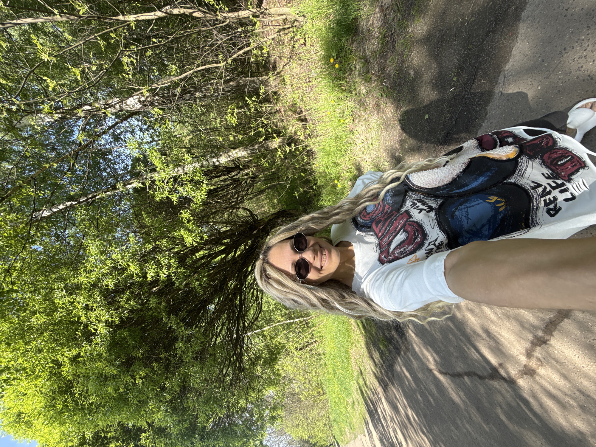 A person with long, blond hair and sunglasses smiles at the camera while standing on a dirt path in Rusanovo, Russia. The person is wearing a white t-shirt with a design on it and white sandals. To the left of the path is a grassy area with trees. The image is taken in daylight.