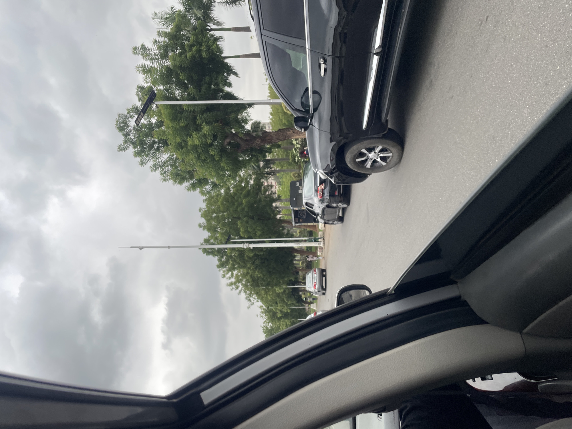 The image depicts a view from inside a vehicle, possibly a car, looking out onto a street in Abuja, Nigeria. The foreground includes elements of the car's interior, such as the roof and side panels. Outside, a line of black vehicles, including SUVs, is visible. The road appears to be paved and the weather overcast, with a grey sky. Trees line the street, interspersed with streetlights. Other cars are further down the street. No people are clearly visible in the image outside of the car interior. The street setting is most likely a residential or commercial area in Abuja.