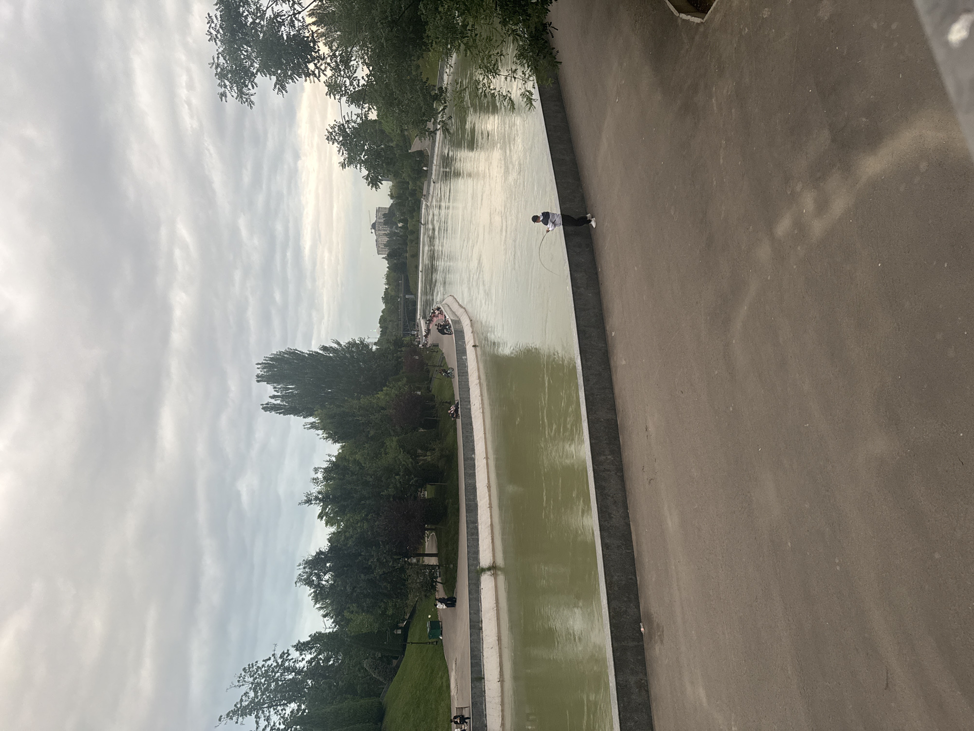 The image shows a long, narrow body of water adjacent to a wide, gray sidewalk. Trees line the left side of the water feature, while the sidewalk occupies the right. A person stands on the sidewalk fishing with a long rod. Other individuals can be seen along the left side of the water, both walking and sitting. The sky is overcast. The location is the Anhor Canal in Tashkent, Uzbekistan.