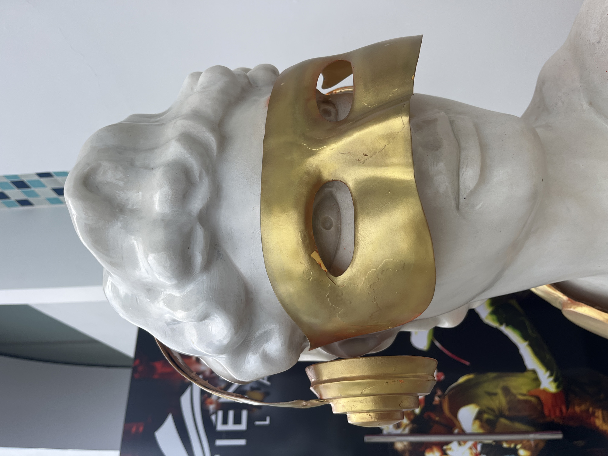 A white plaster bust of a person, adorned with a gold masquerade mask covering the eyes and a gold pair of headphones, is displayed in a public space. The bust is positioned in front of a dark background with text that appears to read "TIEN LA", suggesting it may be an art installation or advertisement for a business or event in Nha Trang, Vietnam. The lighting is bright, and the background shows some architectural elements, including a wall with a geometric pattern. The mask has two eyeholes, revealing parts of the bust's sculpted eyes. The headphones are detailed with stacked discs and rest on top of the bust's head, with one earcup visible on the left side. The overall impression is that of a modern artistic interpretation of a classical sculpture, potentially placed in a commercial or entertainment venue.