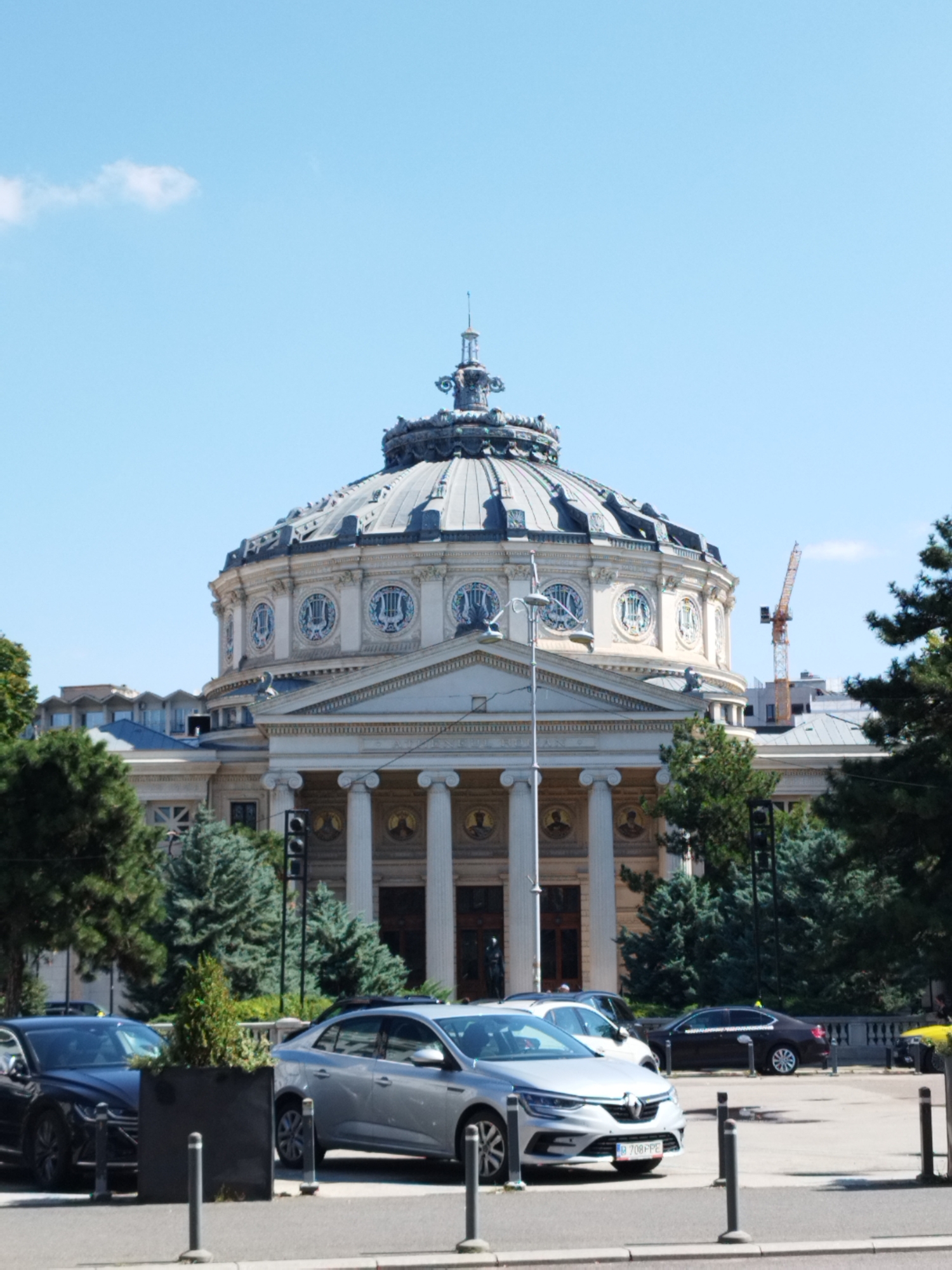 The image shows the Romanian Athenaeum in Bucharest, Romania. The iconic building, with its grand dome and neoclassical facade, is the central subject. In front of the building, several cars are parked on the street, including a silver Renault Megane and a black sedan. Trees and greenery surround the building and line the street. In the background, a construction crane is visible, suggesting ongoing development in the city. The sky is clear and blue, with a few scattered clouds. The overall scene depicts a sunny day in a public urban space.