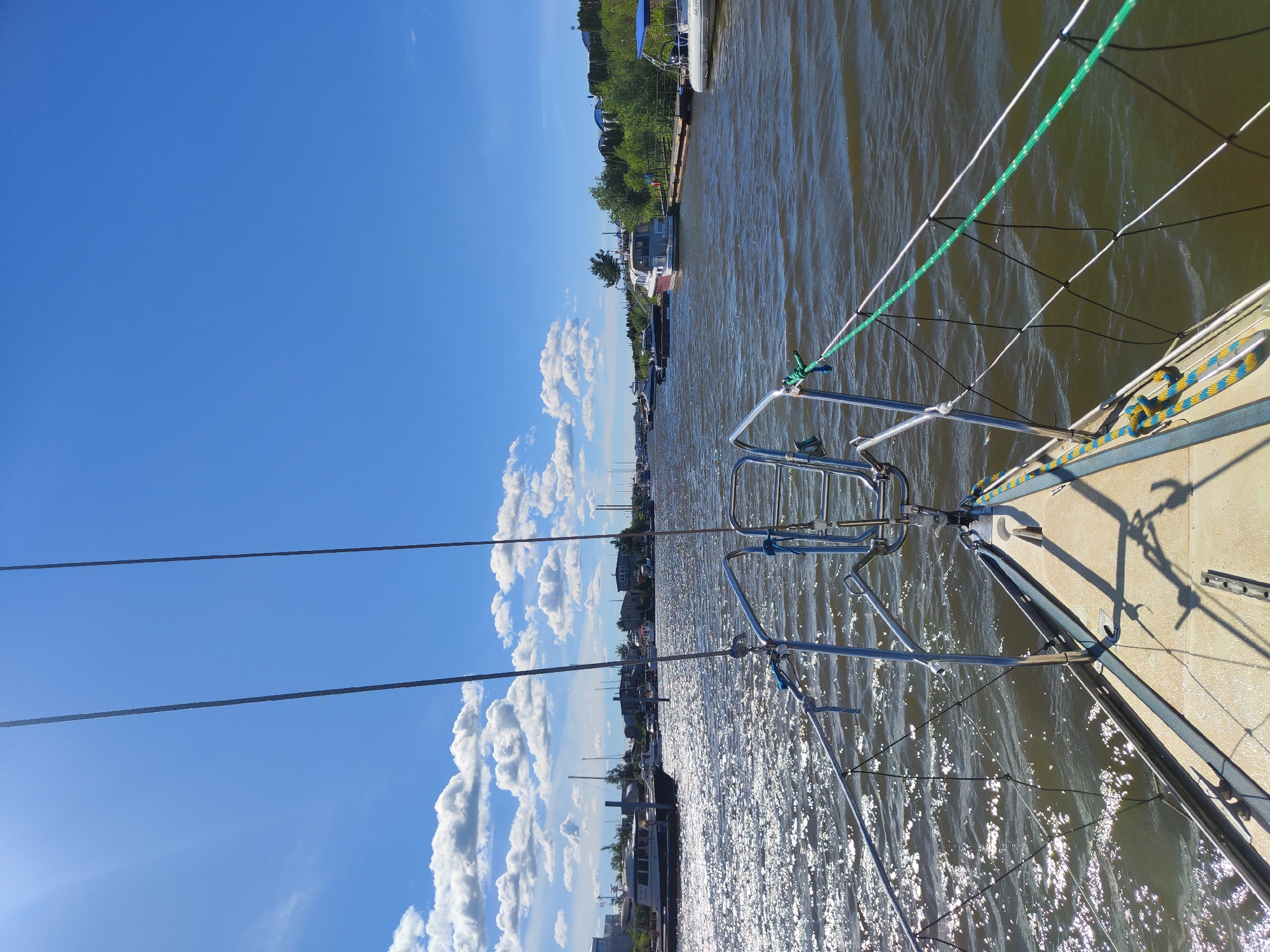 This is a landscape photograph, taken from the deck of a boat, showing a canal or river lined with trees and buildings on the left bank. The water is calm and reflects the bright sunlight. On the right side of the image, the railing and deck of the boat are visible, along with several ropes. In the distance, other boats and structures can be seen along the waterway, suggesting a harbor or a populated waterfront. The sky is a clear, vibrant blue with scattered white clouds, indicating fair weather. The angle of the sun suggests it is either late morning or early afternoon. The presence of boats and the canal setting points to a location that is either a port city or a region with significant waterways. Given the location context of Saint Petersburg, Russia, this scene likely depicts a canal or river within or near the city, possibly during a warm season. There is no visible text in the image.