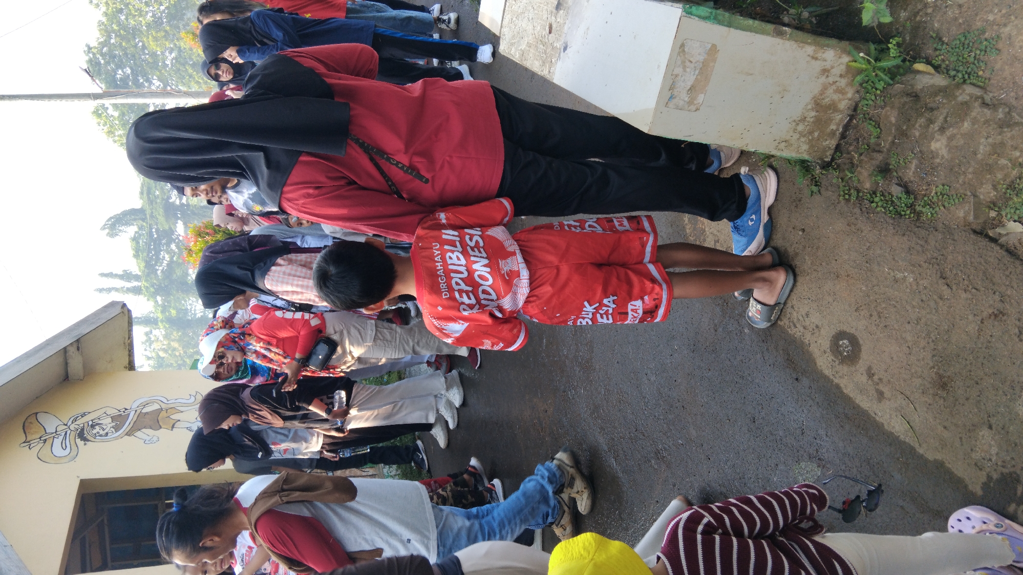 In the city of Posong, Indonesia, a group of people are gathered outside a building. The building has a light-colored exterior with a mural of a cartoonish character holding a flag. In the foreground, a young boy wearing a red t-shirt and shorts with the text "DIRGAHAYU REPUBLIK INDONESIA" is standing. He is wearing grey sandals. To his right, a person wearing a black headscarf and a red shirt stands, their back mostly to the camera. Other people, many wearing headscarves, are visible in the background, some looking towards the left of the frame. The ground is asphalt, and there is some vegetation on the right side of the image. The overall scene appears to be an outdoor gathering.