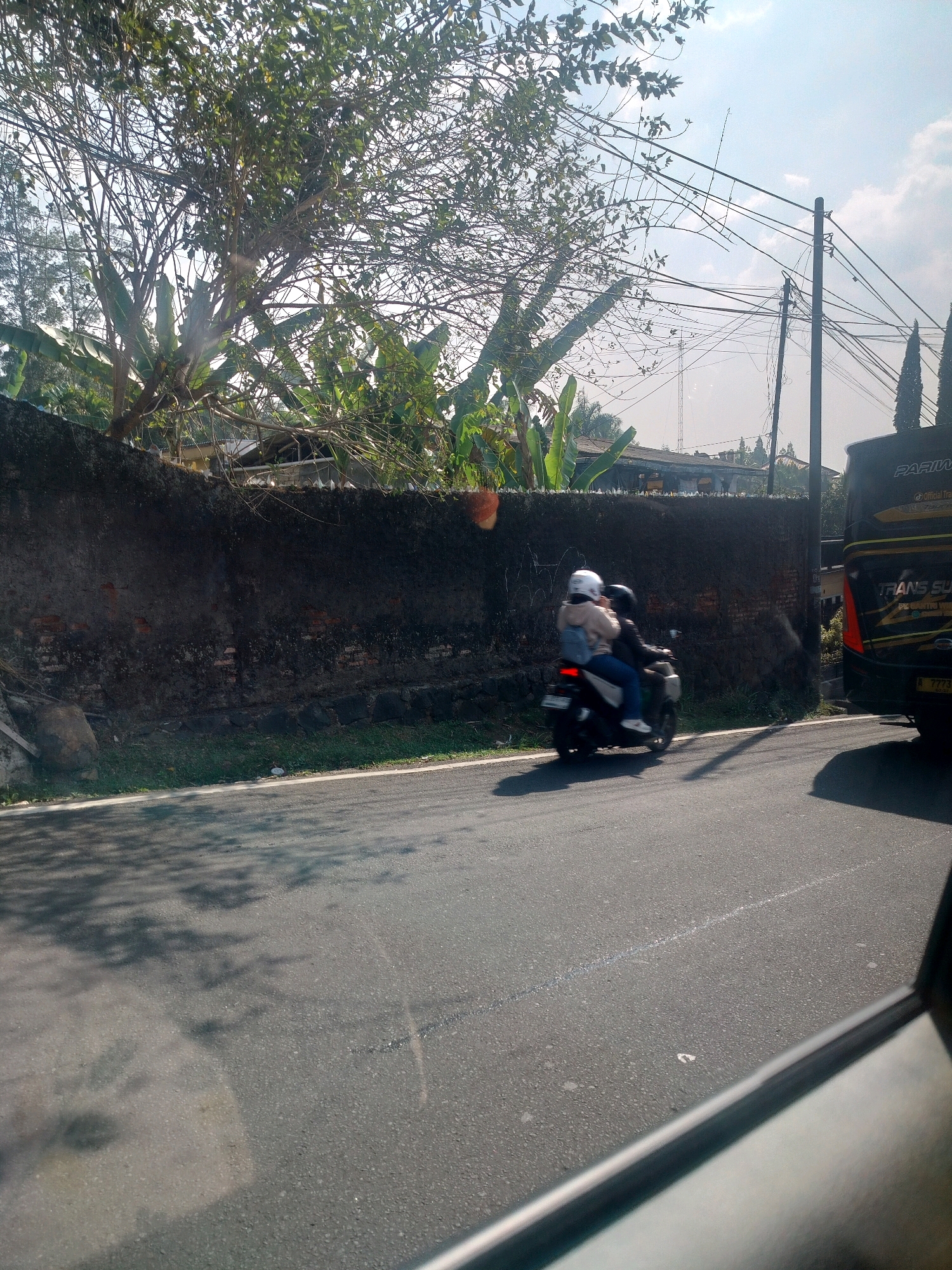 In Cisarua, Indonesia, a motorcycle with two occupants, a driver and a passenger wearing helmets, travels on a paved road. The motorcycle is in motion, heading away from the viewer. Behind the motorcycle, a black bus with yellow accents is visible. The environment features a weathered, dark-colored stone wall bordering the road, with lush green vegetation and trees above it. Power lines crisscross the sky, supported by utility poles. The sky is partly cloudy with bright sunlight casting shadows on the road.