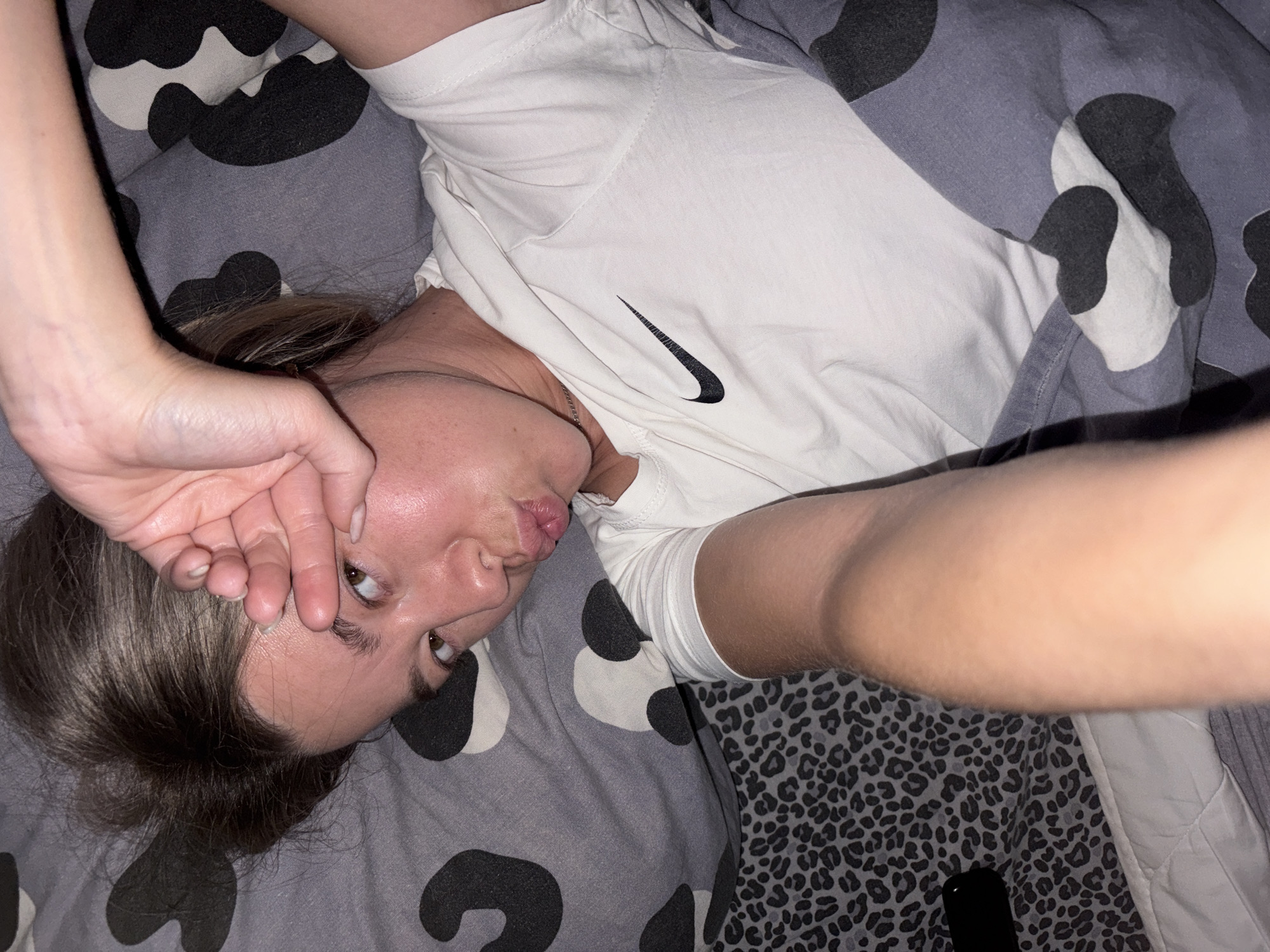 The image shows a young woman lying in bed. She is wearing a white Nike t-shirt and is partially covered by a grey blanket with a black and white cow print pattern. Her left hand is covering her eye, and she is puckering her lips. The background is a bed with patterned sheets and a pillow. The location is not specified in the image or accompanying text.