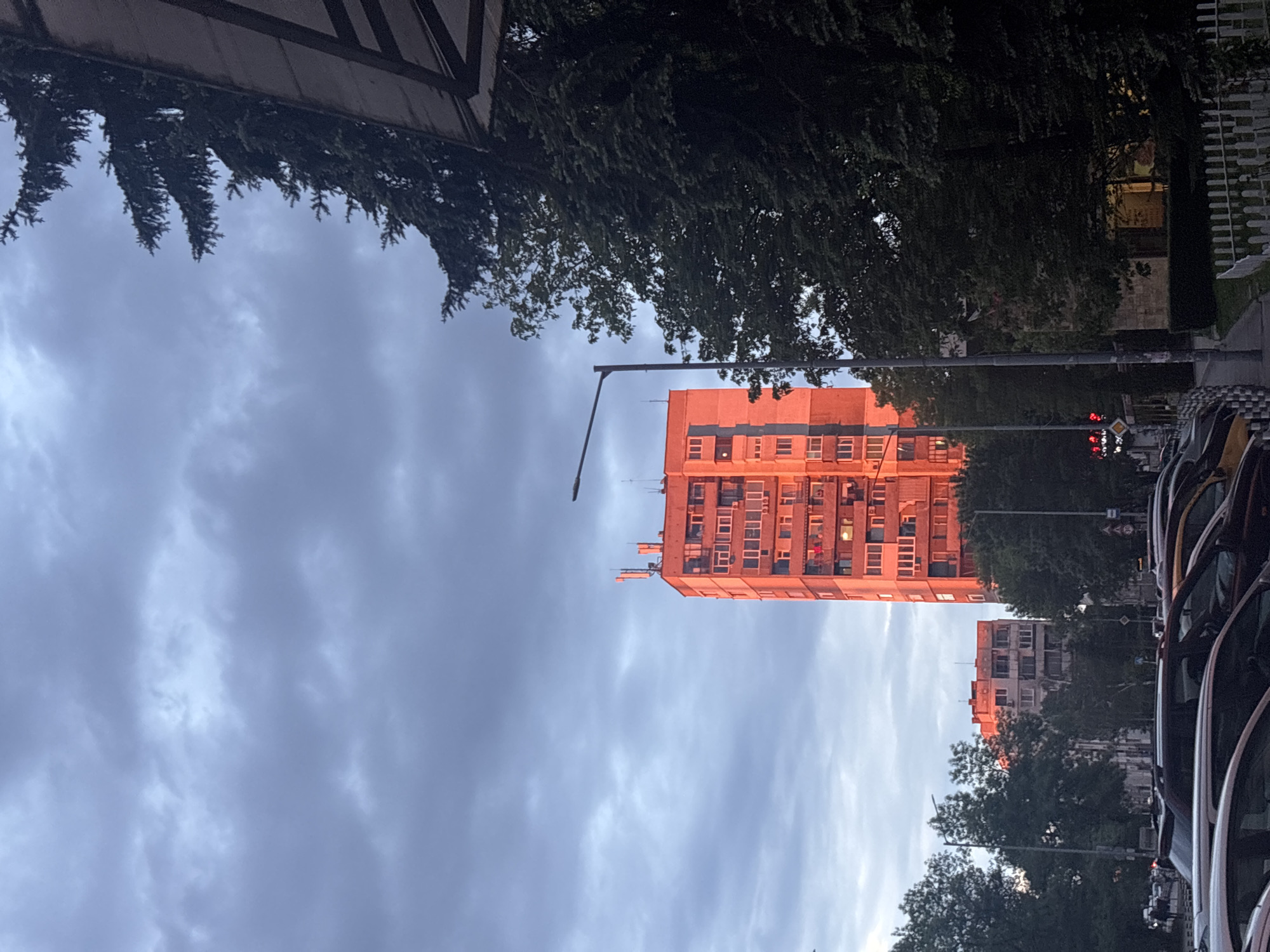 The image captures a view in an urban setting in Unknown, Unknown. In the foreground on the right, a row of vehicles is visible, partially obscuring the view. Beyond the vehicles, a street with traffic lights is present. The main focus is a tall residential building in the center, illuminated with a warm orange glow, possibly from the setting sun. Behind this building, another, smaller building is visible. Trees frame the buildings on both sides. The sky is overcast with grey clouds. A streetlight extends upwards from the ground towards the left of the building.
