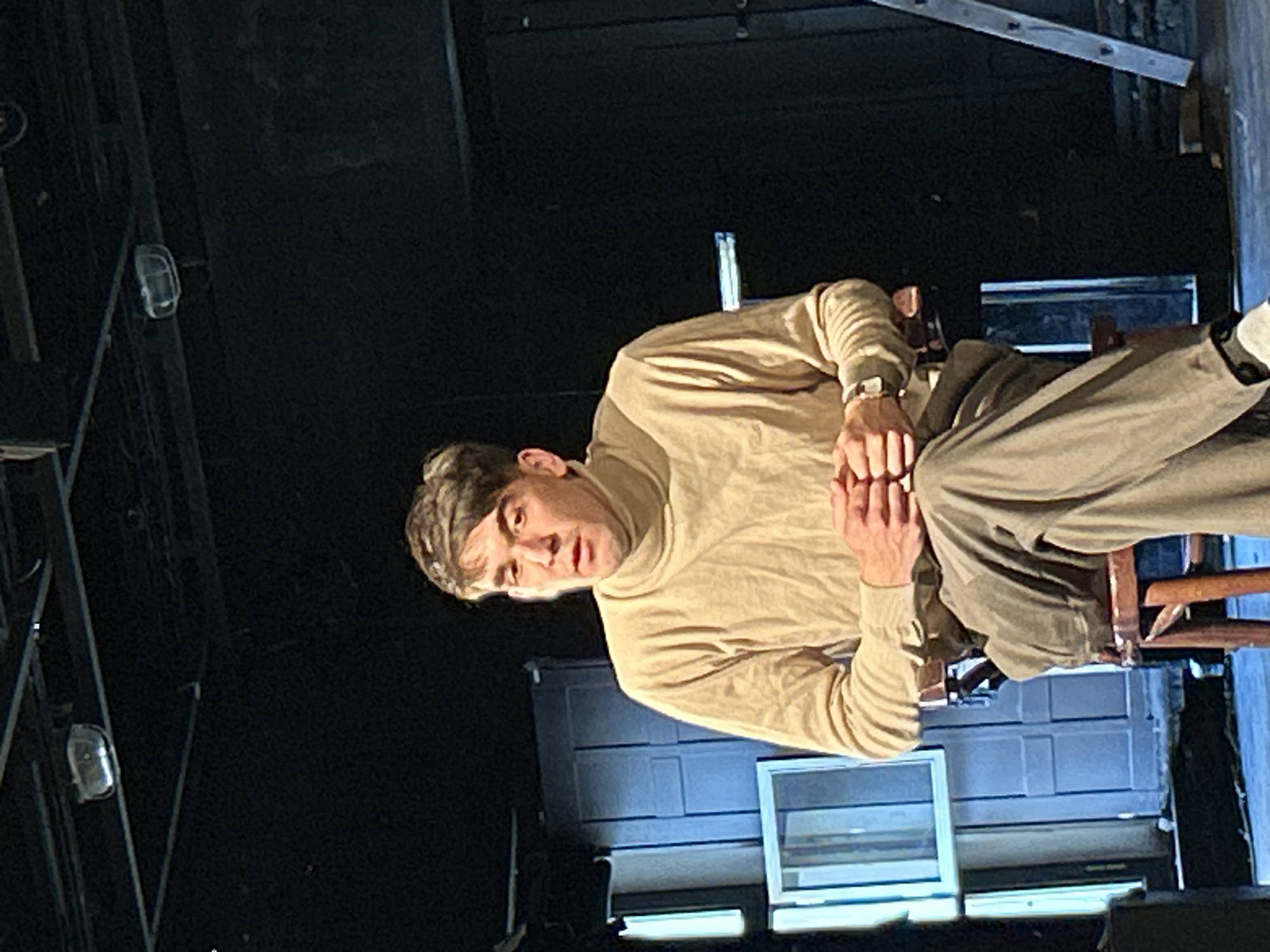 A man is depicted seated in what appears to be an indoor, dimly lit environment, possibly a stage or a studio set. He is positioned slightly off-center, facing forward, and looking directly at the viewer with a serious or contemplative expression. He is wearing a light tan or beige long-sleeved turtleneck shirt and olive green trousers. His hands are clasped together in front of him, resting between his knees. He has short, dark brown hair. He is seated on a dark wooden chair, parts of which are visible beneath and behind him.
The background is largely dark, with areas illuminated by specific lighting. To the man's right, there is a structure resembling a wall with horizontal paneling, and below it, a section with a horizontal, reflective surface that could be a window or a screen. To his left, the background is darker, but some structural elements, possibly wooden beams or stage rigging, are discernible. A small, clear, oval-shaped light fixture is visible mounted on a vertical support in the upper left background. The lighting in the scene is focused on the man, creating a distinct contrast with the shadowed background, suggesting a deliberate and controlled lighting setup. There is no visible text in the image. The scene conveys an intimate and somewhat dramatic atmosphere, reminiscent of a theatrical performance, rehearsal, or a carefully staged portrait.
