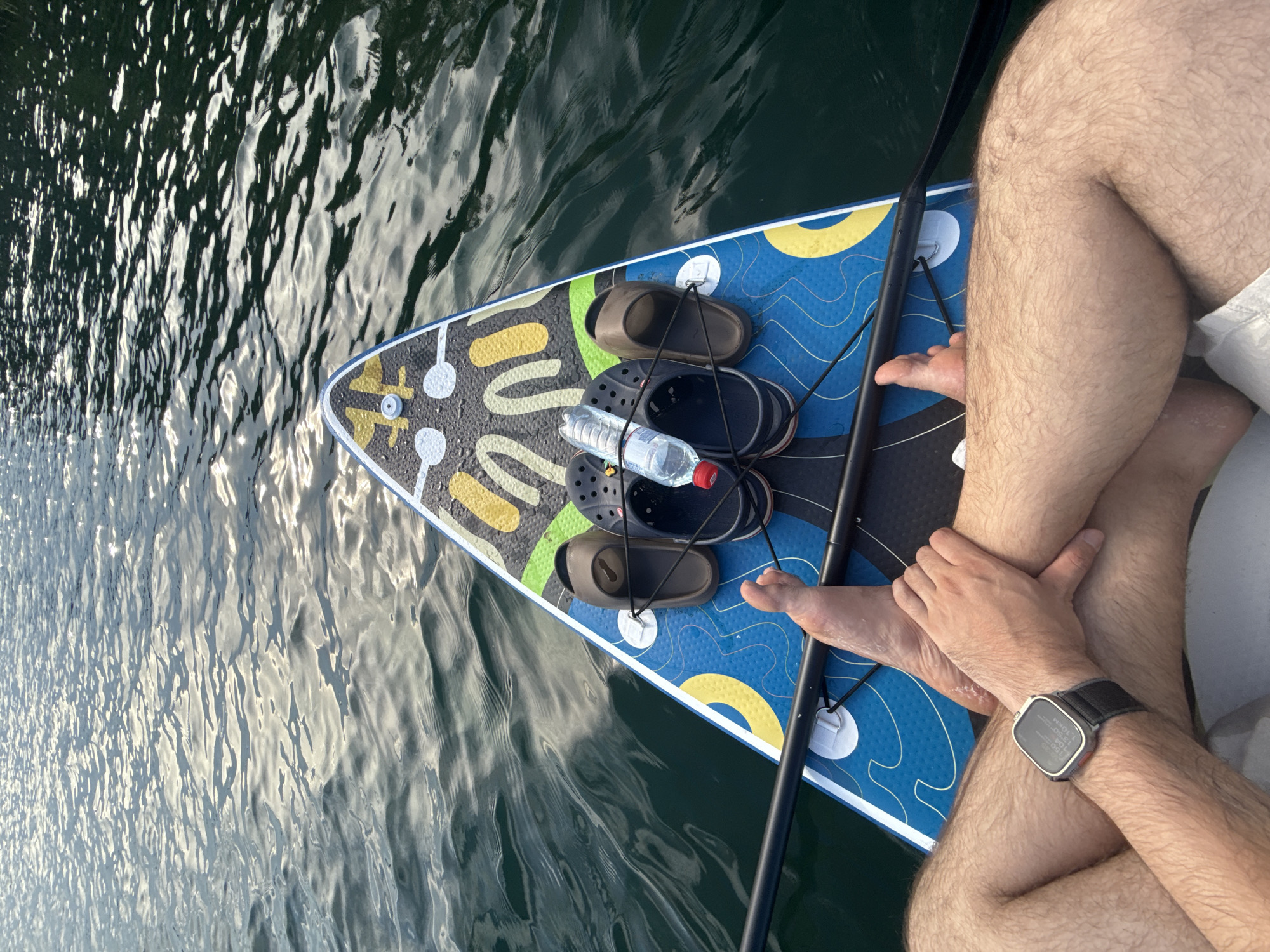 The image shows a person sitting on a paddleboard in the water. The person is wearing white shorts and a watch on their left wrist. Their legs are crossed. On the paddleboard, there are two pairs of sandals, a bottle of water, and a paddle. The water is dark and reflects the sky, indicating it is likely daytime. The scene appears to be a calm body of water, possibly a lake or calm sea, given the rippling surface. The overall impression is one of relaxation and outdoor activity. The watch on the person's wrist shows the time as 11:53. The location is Zaosinovo, Russia.