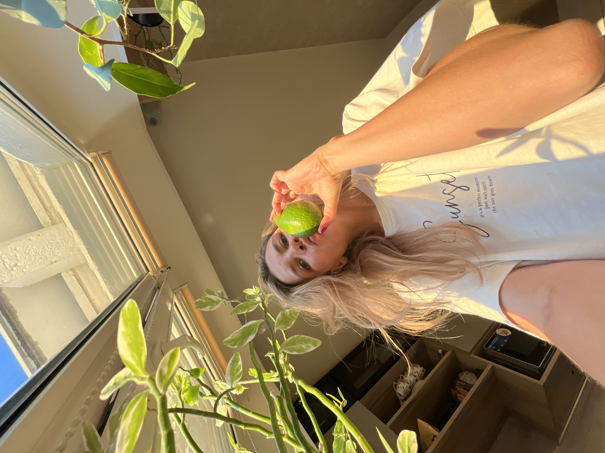 A woman is holding a lime in front of her face, near a skylight. She is wearing a white t-shirt with text on it. The room appears to be a home interior with shelves and plants visible. There is a skylight overhead with sunlight streaming through it, casting shadows. The location is not identifiable as Chaikovsky, Russia.