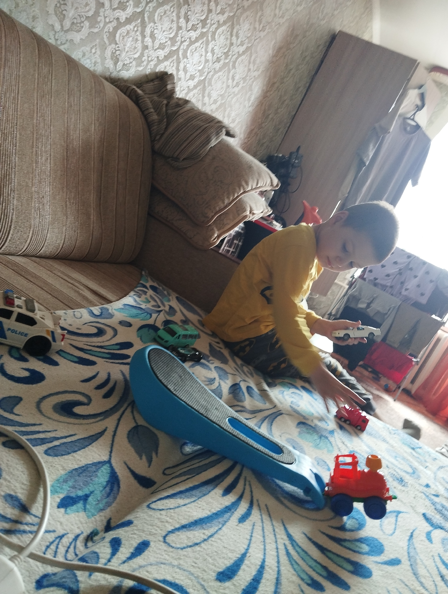 In an indoor setting, likely a bedroom or living room, a young boy with short brown hair, wearing a yellow long-sleeved shirt and patterned pants, is seen on a bed with a blue and white patterned comforter. He is holding a small white toy car. To his left, a toy police car with "POLICE" written on its side is visible on the comforter. A small green toy vehicle and a tiny black toy car are also present. A blue toy, possibly a scooter or sled, is positioned on the comforter in the foreground. In the background, a couch with striped cushions is visible. A wardrobe with hanging clothes and some toys, including a red toy truck and a bright orange toy, are also present. The overall scene depicts a child playing with toys in a domestic environment. The provided information about the location being Kingisepp, Russia, cannot be verified from the image alone.