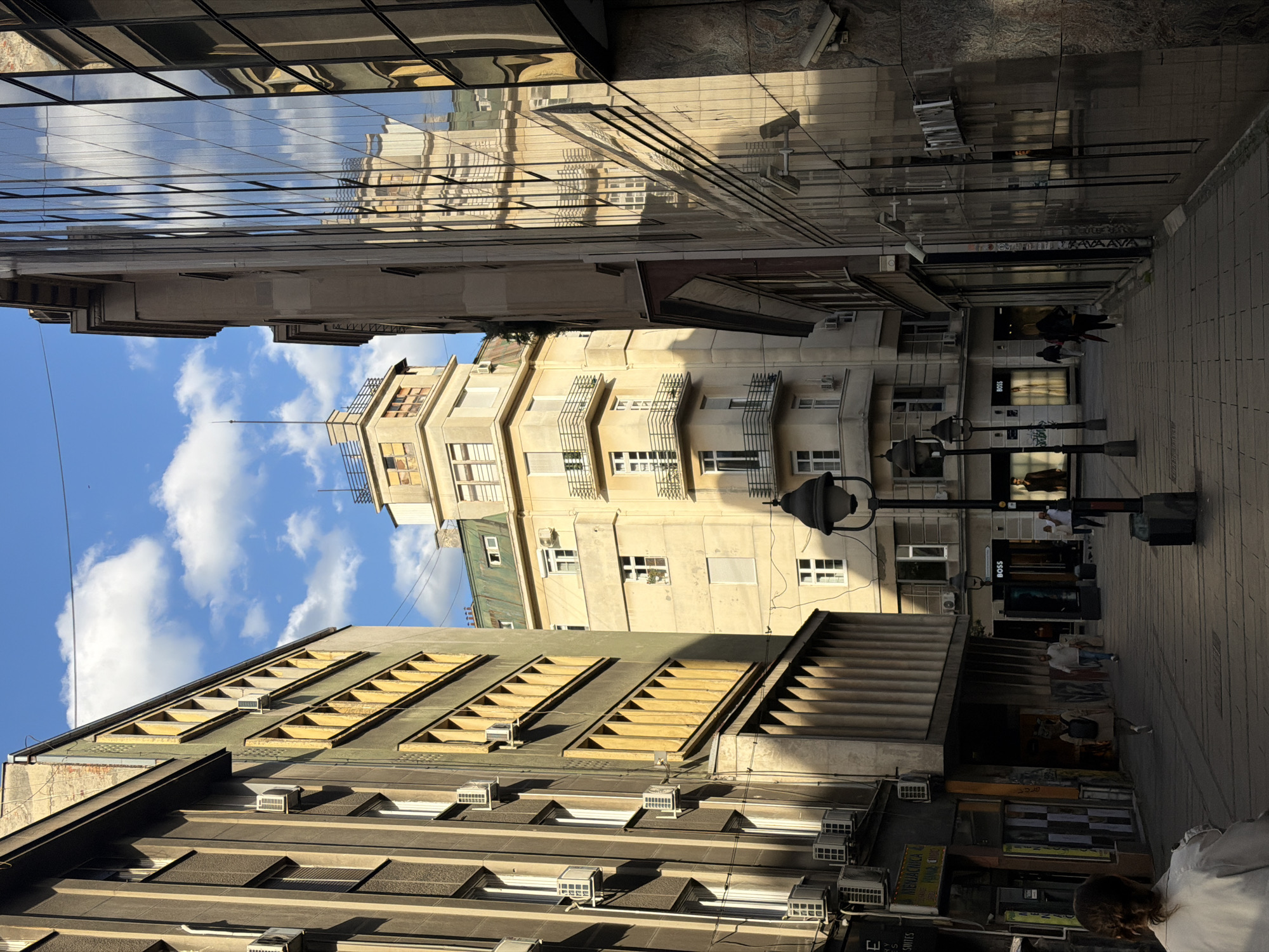 The image depicts an alley in Belgrade, Serbia, lined with various buildings. The sky is visible with scattered clouds. The street is paved and contains street lamps and pedestrians walking in both directions. Buildings display different architectural styles, some with balconies, windows, and air conditioning units. One building has a modern reflective glass facade that mirrors other buildings and the sky. A store displays a "BOSS" sign. The overall scene suggests an urban environment with a mix of historical and modern architecture.