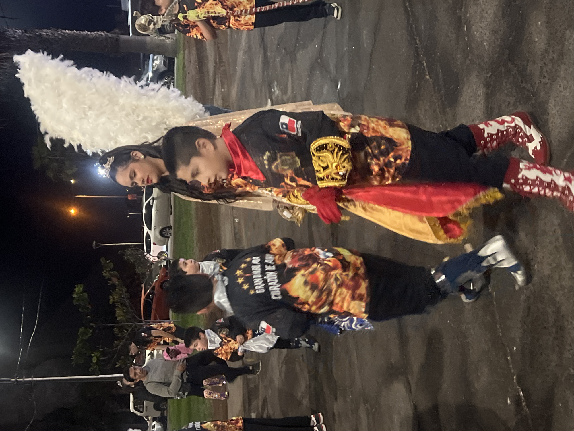 In the city of Arica, Chile, at night, a group of people are gathered outdoors. A young boy in the foreground wears a black costume with vibrant orange and red designs, a red bandana, and elaborate gold gauntlets on his hands. He is wearing red and white patterned boots. Behind him, another young boy in a similar costume is visible. To the left, a woman with dark hair, wearing a crown, has large white wings attached to her back, suggesting she is dressed as an angel. She is wearing a shimmering gold dress. Other individuals in similar costumes are in the background, some holding objects with flame-like patterns. Cars are parked in the background, and streetlights illuminate the scene.