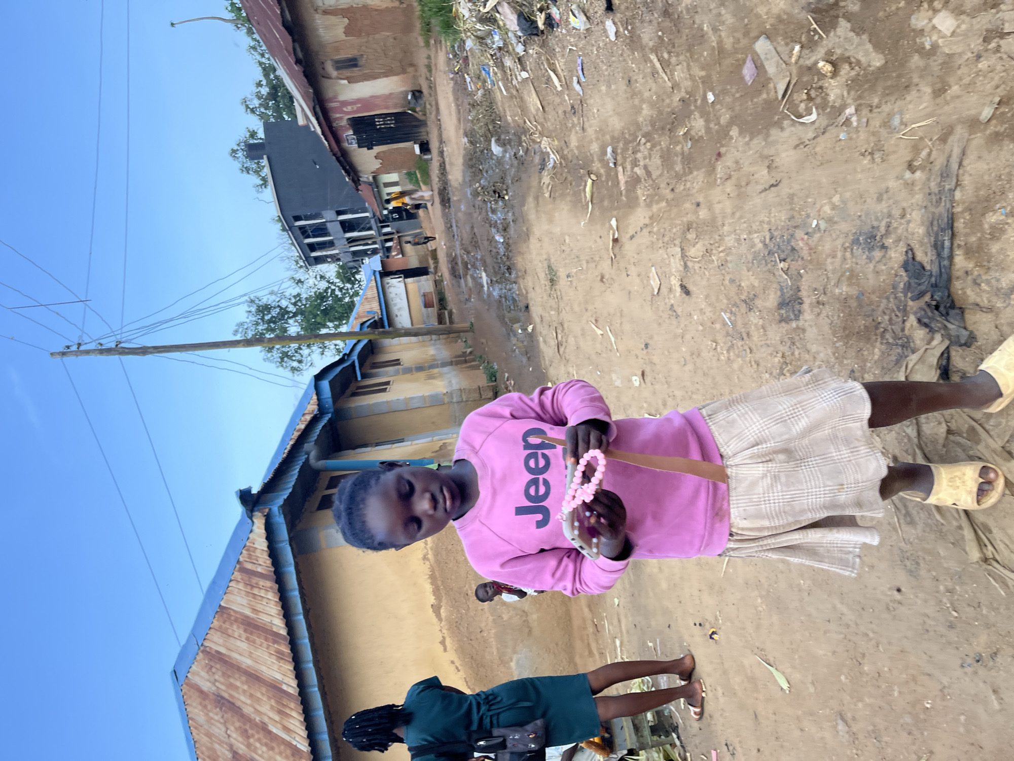 In Rafin Par, Nigeria, a young girl stands in a dirt path, wearing a pink sweatshirt with "Jeep" printed in large black letters and a plaid skirt. She holds a pink beaded necklace and a cell phone. Behind her, another girl in a green dress and a smaller child are visible. The environment is characterized by a dirt road with debris, mud puddles, and simple buildings with corrugated metal roofs. Power lines crisscross the sky above.