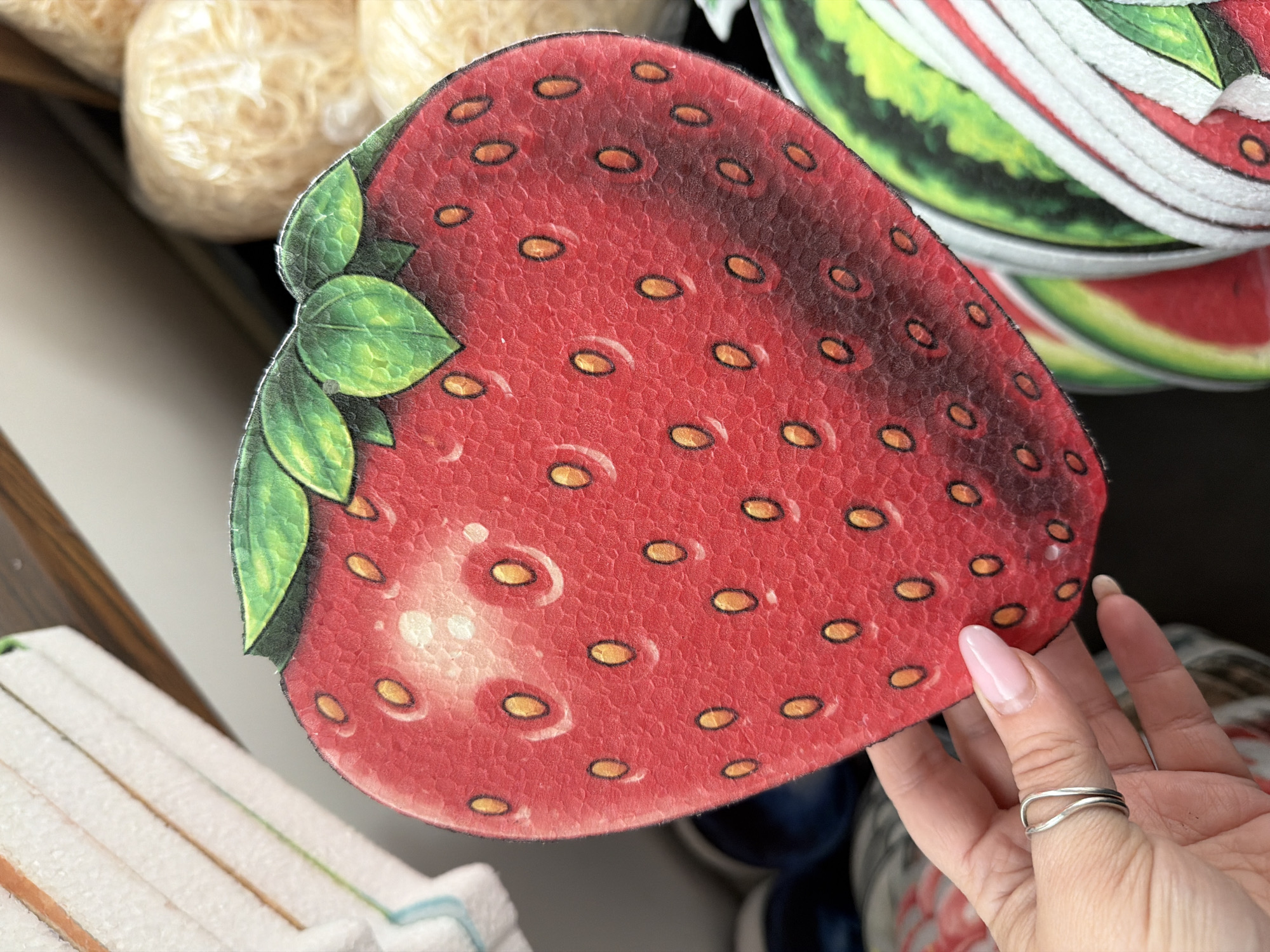 A close-up shot focuses on a person's hand holding a large, red, strawberry-shaped decorative item. The strawberry has green leaves at the top and numerous small, yellow seeds with black outlines scattered across its surface. The item appears to be made of a textured material, possibly felt or foam, with a printed design. The hand holding the strawberry has light-colored skin and a silver ring on the index finger. The fingernails are painted a light pink color. In the background, out of focus, are other decorative items, including what appears to be a stack of white, striped objects on the left and a round, light-colored object wrapped in plastic on the right. Further in the background, parts of other fruit-shaped decorations, possibly watermelons, are visible. The image does not contain any visual information that can be used to determine the location within the city of Ternopil, Ukraine.