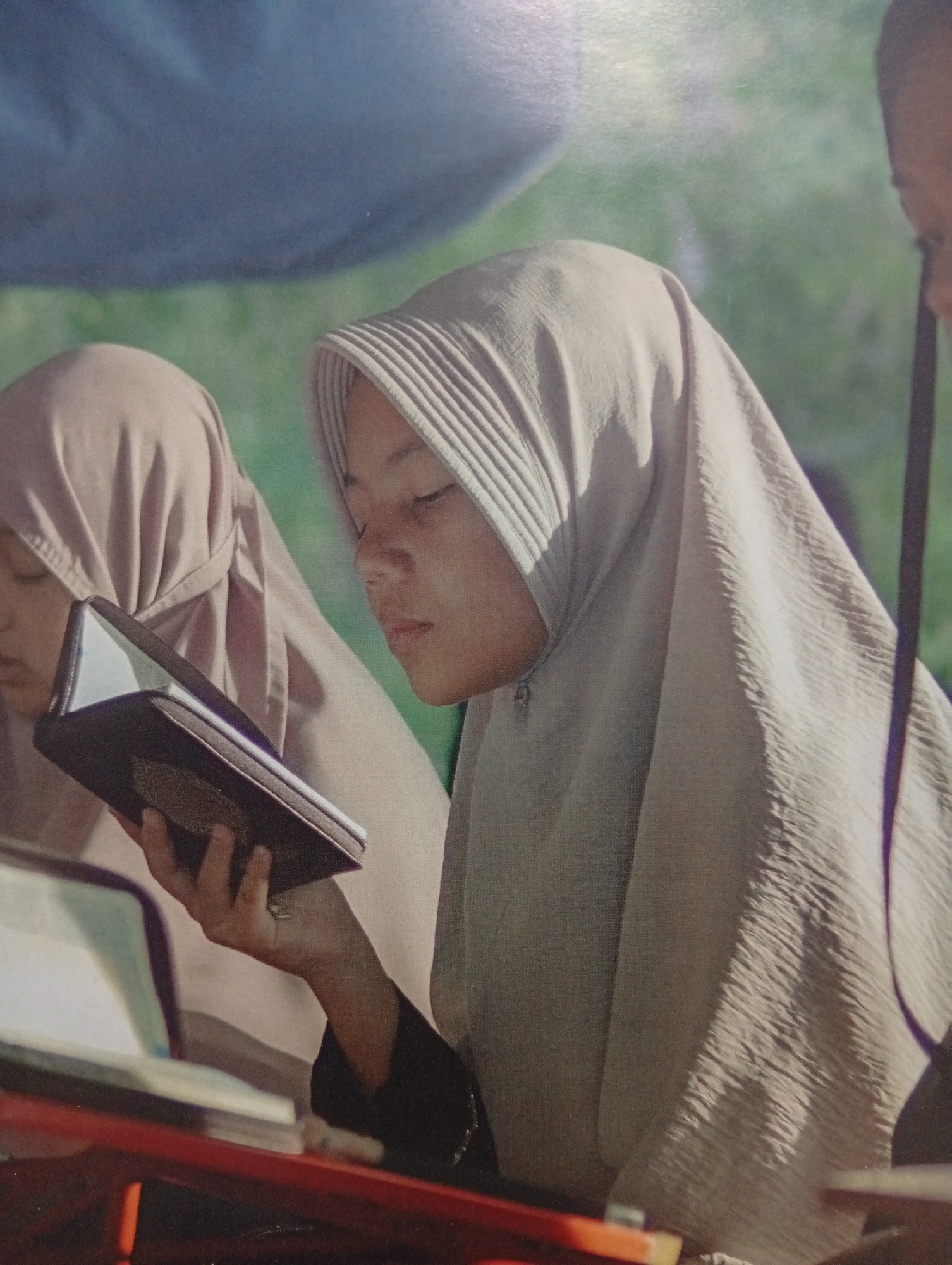 In Tlogomulyo, Indonesia, two young women wearing hijabs are engrossed in reading. The woman in the foreground, positioned slightly to the right, wears a light-colored hijab with subtle pleating around the forehead. She is holding open a dark-covered book, possibly a Quran, with her left hand and her gaze is directed downwards at the pages. Her right hand is also visible, resting on the book. To her left and slightly behind, another woman in a similarly colored hijab, though with a smoother texture, is also reading. She is looking down at a book or object that is not entirely visible. Both women appear to be seated, with a red surface in the lower part of the frame suggesting a table or desk. The background is softly blurred, with hints of green foliage and a dark blue element, possibly a canopy or sky. The lighting suggests an outdoor or well-lit indoor setting.