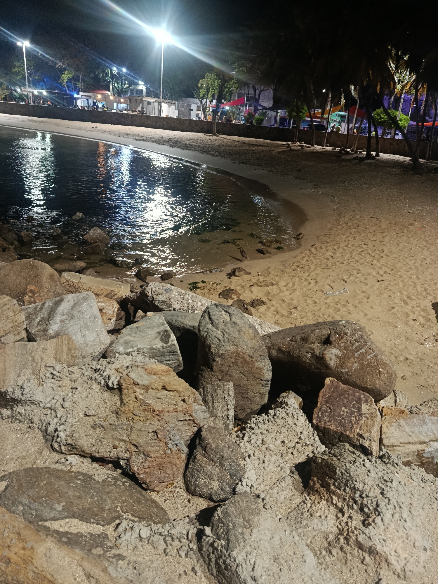The image shows a night scene at a beach in Juan Griego, Venezuela. In the foreground, there is a pile of large, weathered rocks. Beyond the rocks, there's a sandy beach that slopes down to the water's edge. The water reflects the light from street lamps and nearby buildings, creating a shimmering effect. On the far side of the beach, there's a raised area with buildings and trees, indicating an urban setting. There are people and some structures visible in the background suggesting a public space or gathering area. The image is taken at night, with the primary light sources being artificial lighting.