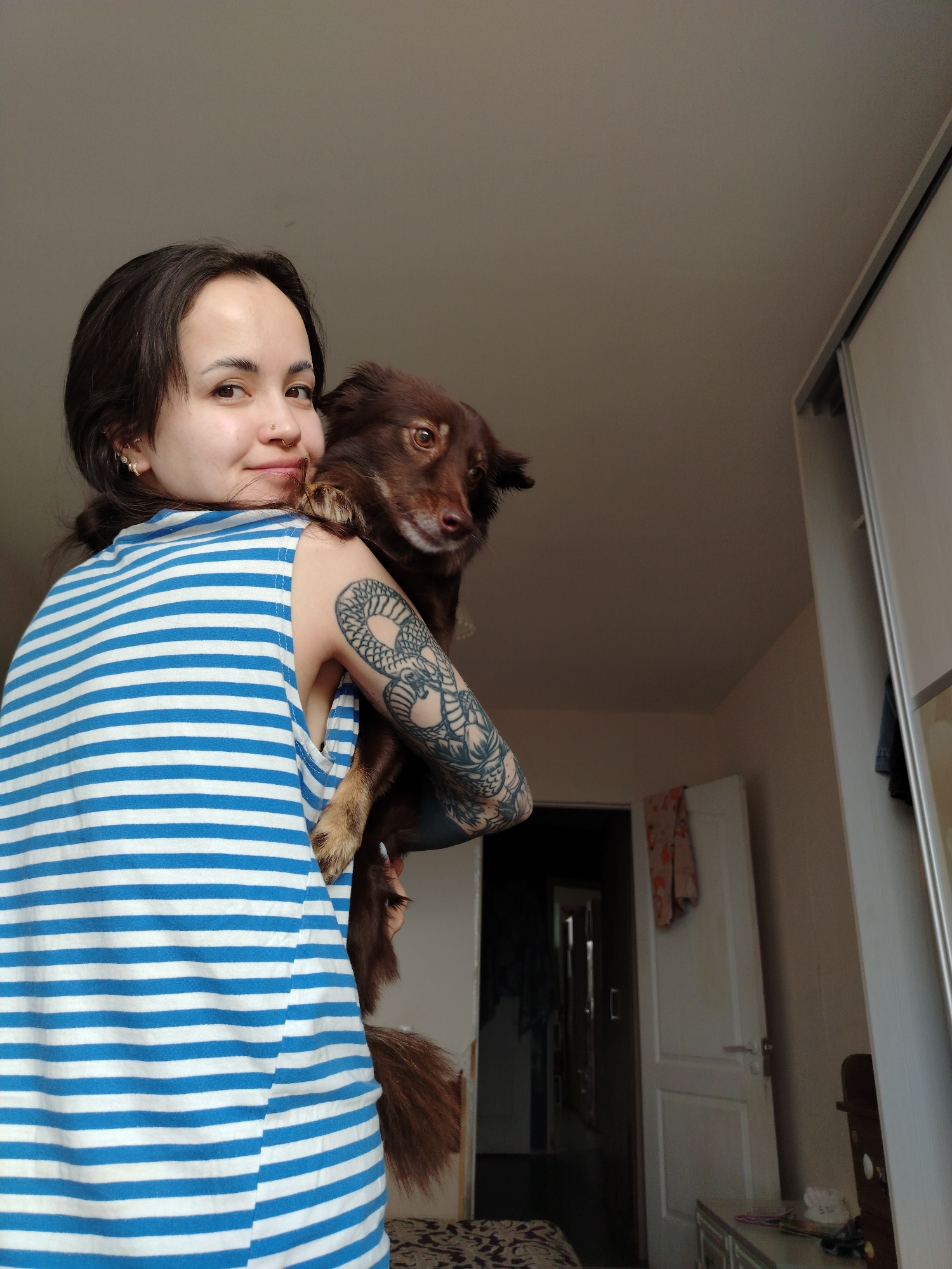 A young woman is captured in a close-up, half-body shot, looking over her left shoulder towards the viewer with a gentle smile. She has dark hair styled in a bun or ponytail, a nose ring, and multiple small hoop earrings. She wears a sleeveless blue and white striped top. Her left arm, featuring a prominent, intricate tattoo depicting what appears to be a serpent or dragon, holds a small to medium-sized dog. The dog has dark brown fur and is looking towards the camera with a slightly curious expression.
The scene takes place indoors, likely a bedroom or living area within a home in Noviy, Russia. The background features light-colored walls. To the right, a white wardrobe with sliding doors is partially visible, alongside a dresser or table with a few small items. Behind the woman's shoulder, a white door stands slightly ajar, revealing a darker hallway beyond with another open doorway. The lighting suggests it is daytime. No text is visible in the image.