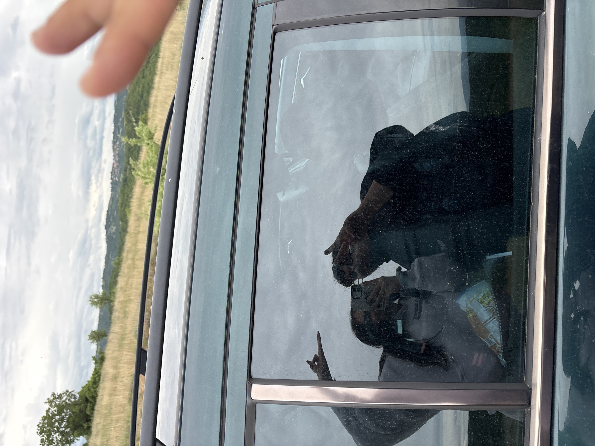 The image shows a reflection in the window of a vehicle. The reflection captures two individuals taking a photo with a smartphone. One person is wearing a dark-colored top, and the other appears to be wearing a grey top. The second person is extending their middle finger. Inside the vehicle, there is an item that looks like a package of chips. The background visible through the window and in the reflection features a landscape of rolling hills and fields with some vegetation. Based on the prompt the location is Marulevo, Bulgaria. A partial hand is visible in the upper-left corner of the image, seemingly belonging to someone outside the vehicle.