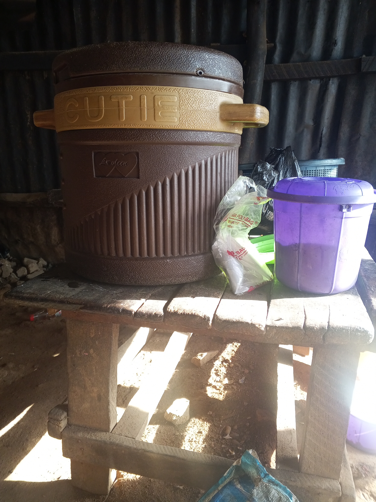 An outdoor scene at ground level shows a large, brown plastic cooler labeled "CUTIE" on a rustic wooden table. To the right of the cooler are a clear plastic bag, a purple plastic bin with a lid, and a grey plastic basket. The table is made of rough-hewn planks supported by wooden posts. The ground beneath the table is covered in dirt and debris, with sunlight casting dramatic shadows. In the background, corrugated metal sheeting forms a wall. The overall impression is one of utilitarian storage in an outdoor, possibly rural or workshop setting.