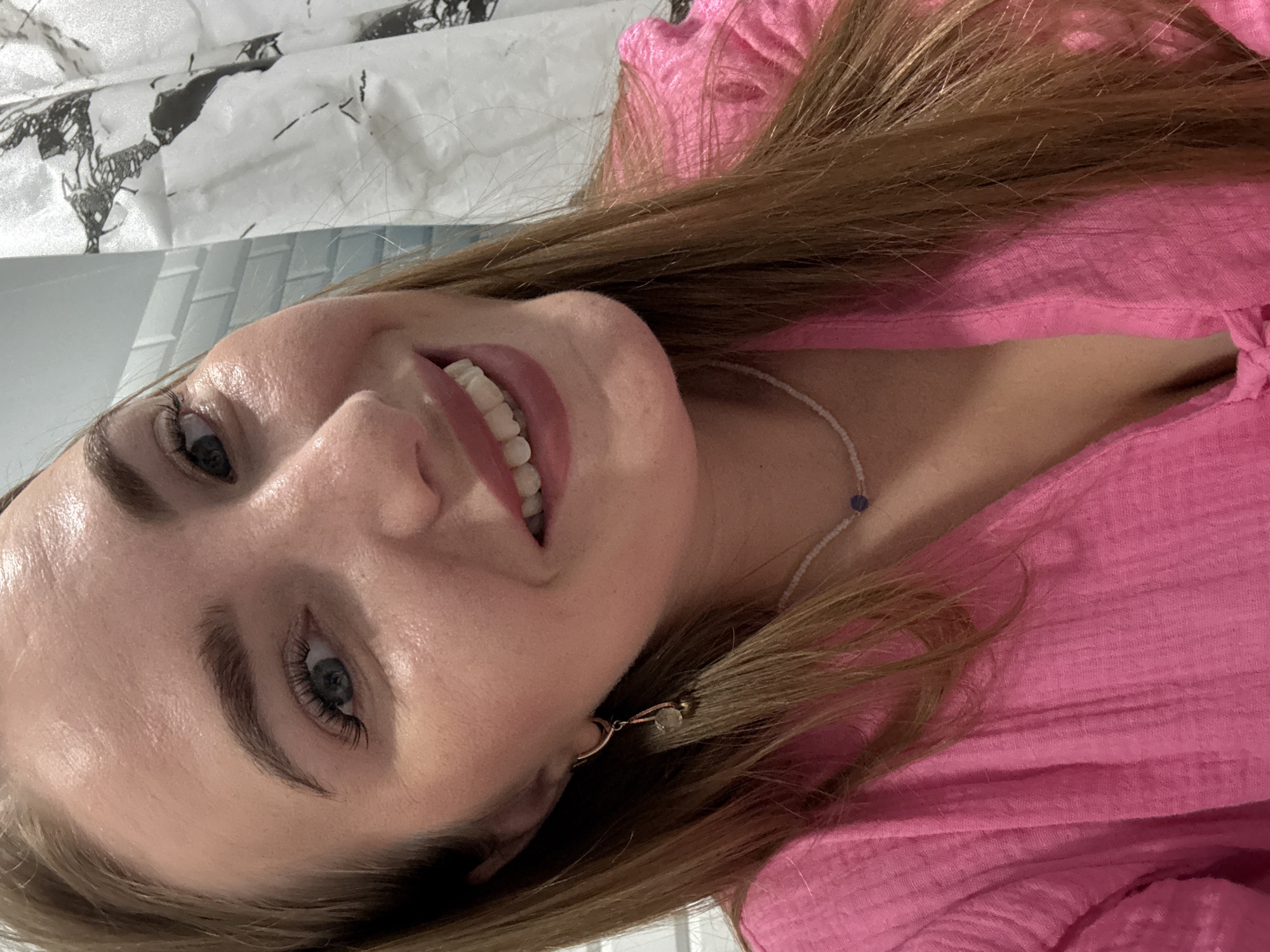 A close-up, head-and-shoulders selfie of a young Caucasian woman with long, light brown hair, fair skin, and blue eyes. She is wearing a bright pink, textured garment, possibly a blouse or dress, with a V-neckline. A delicate beaded necklace with a small blue stone is visible around her neck. She has a subtle smile, showing her teeth. She is wearing a gold hoop earring with a dangling pendant. The background is blurred, but suggests a domestic setting, with tiled wall on the left and a shower curtain with a black and white abstract pattern on the right. There is no indication that this scene is in Kazan, Russia.